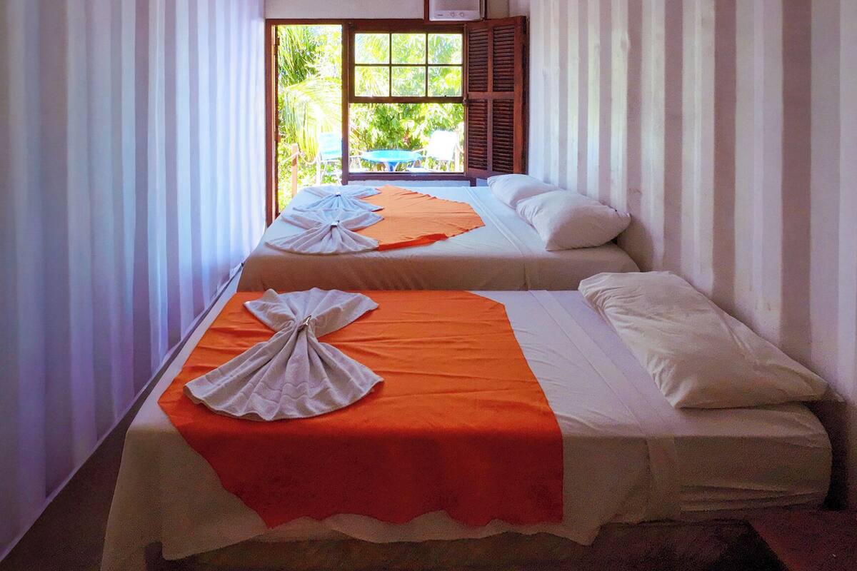 Two single beds with orange and white linens, a window with wooden shutters, and a view of greenery outside.