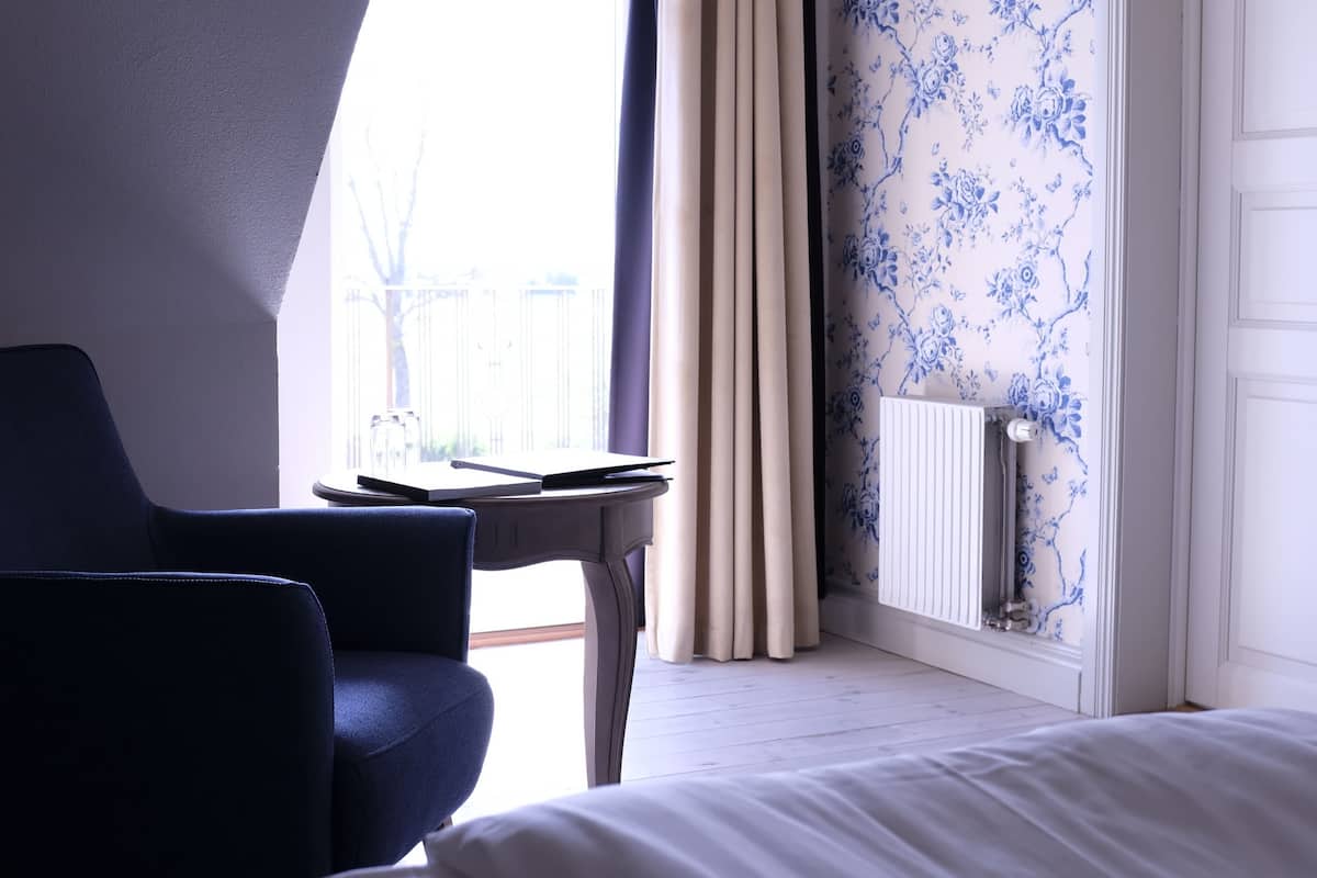 A hotel room with a bed, a chair, a table with a book, curtains, and a radiator.