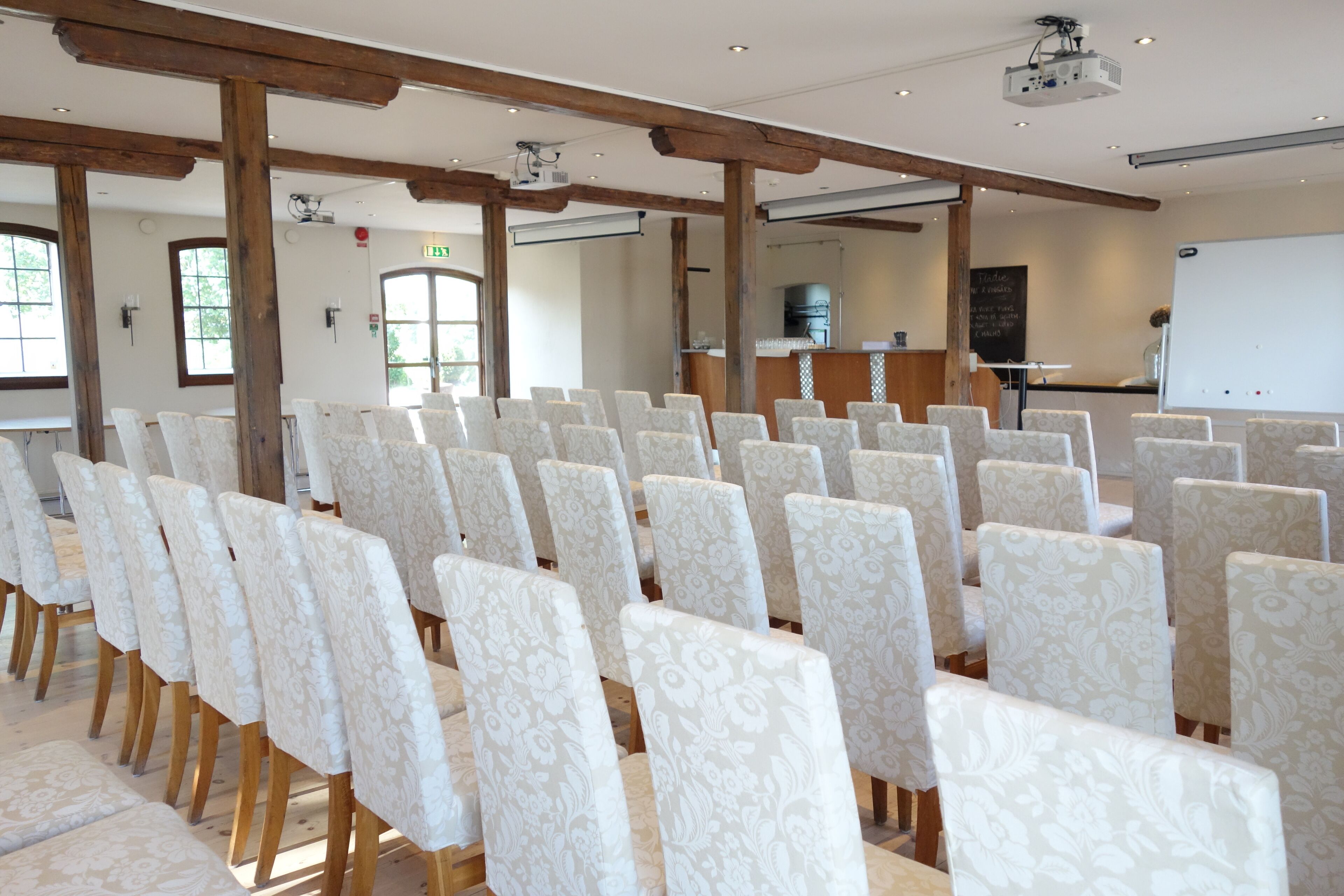 Un salón de conferencias con filas de sillas, vigas de madera, un proyector y una pizarra.