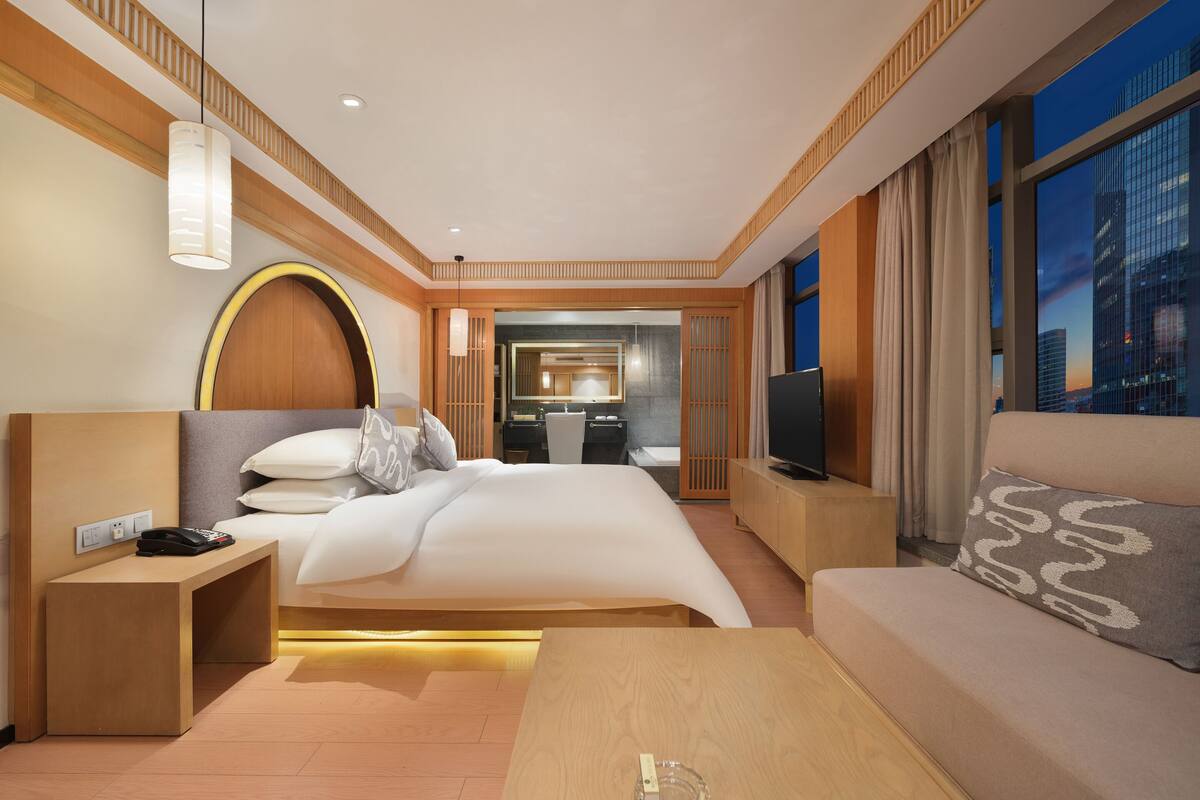 Une chambre d’hôtel moderne, dotée d’un grand lit, d’une télévision, d’un canapé et d’une salle de bain visible à travers une porte ouverte.