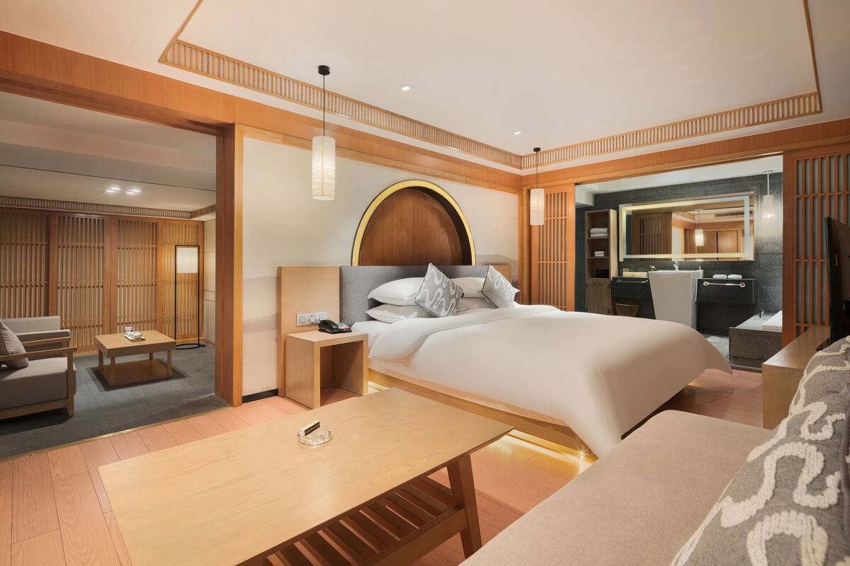 Une chambre d’hôtel moderne dotée d’un grand lit, d’un coin salon avec un canapé et un fauteuil, d’une table basse en bois et d’une salle de bain avec un grand miroir.