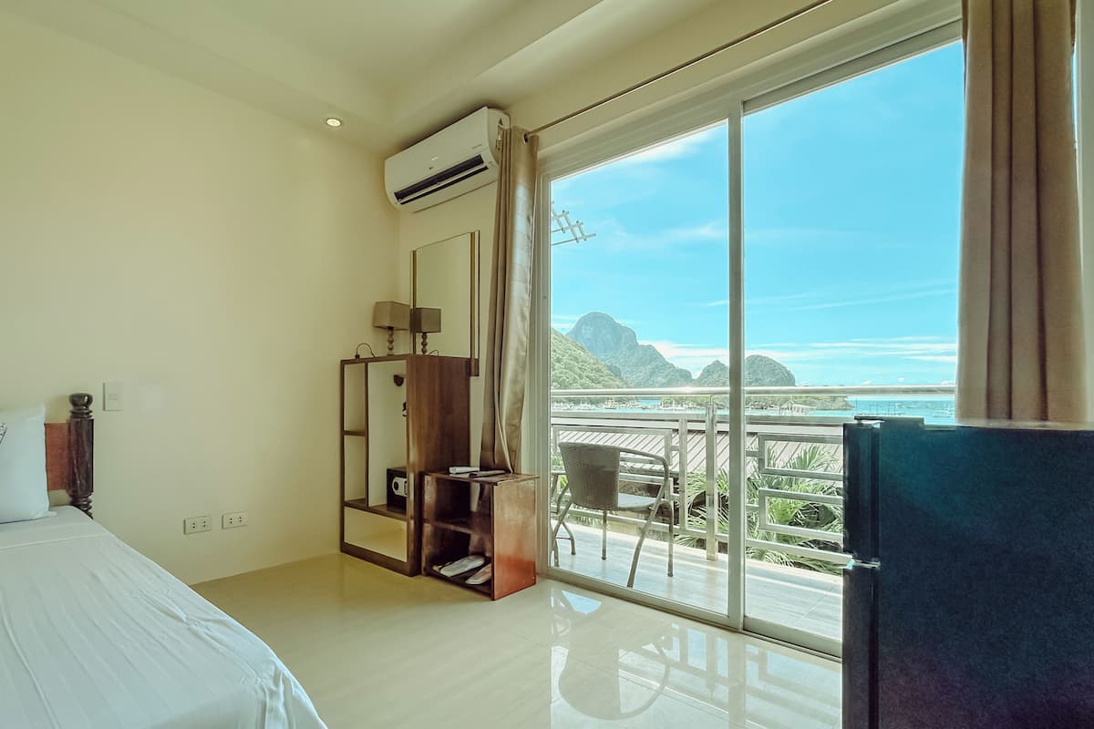 A hotel room with a bed, a desk, a chair, and a large window offering a view of the sea and mountains.