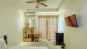Ein Hotelzimmer mit Bett, Deckenventilator, Klimaanlage, Fernseher, kleinem Tisch mit Lampe, Schuhregal und einem Fenster mit Vorhängen.