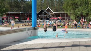 Une zone de piscine où des personnes nagent et se détendent, entourée de parasols et d’un bâtiment à la façade en bois.