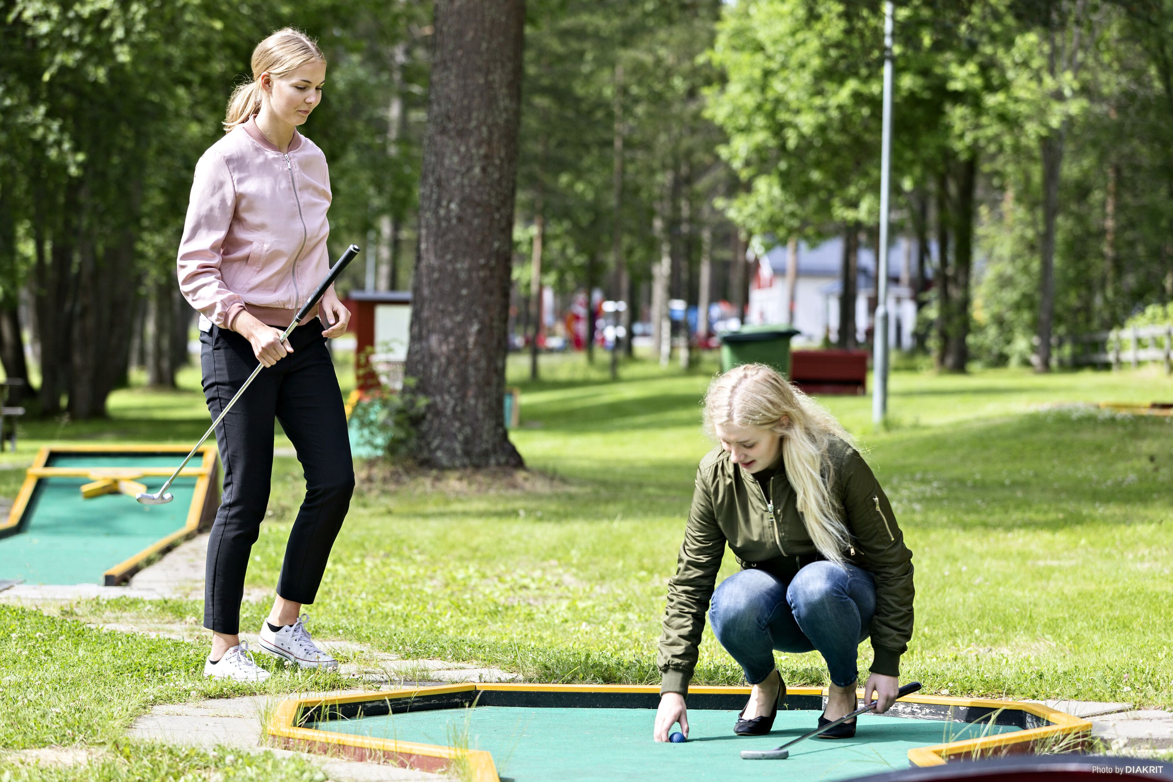 Two people playing miniature golf in an outdoor setting with trees and a green field.