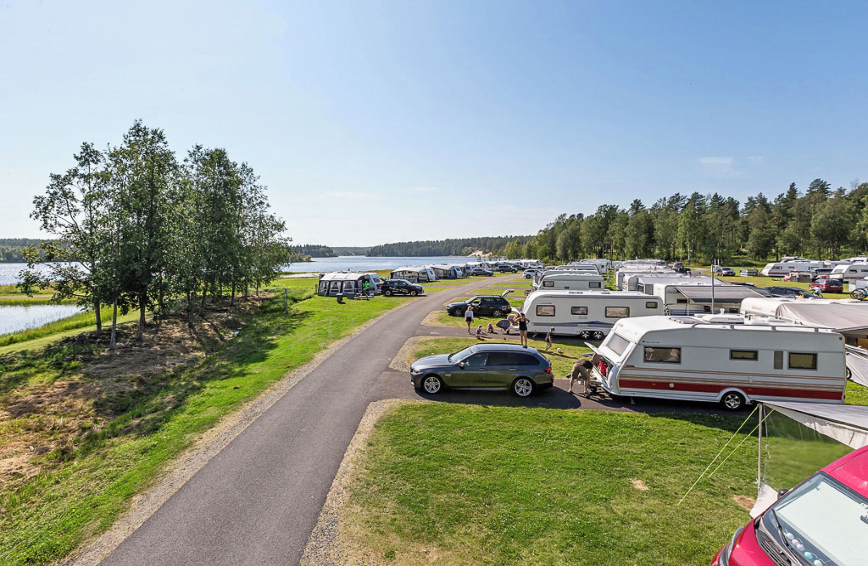A campsite with RVs and cars parked along a road, surrounded by trees and a lake.