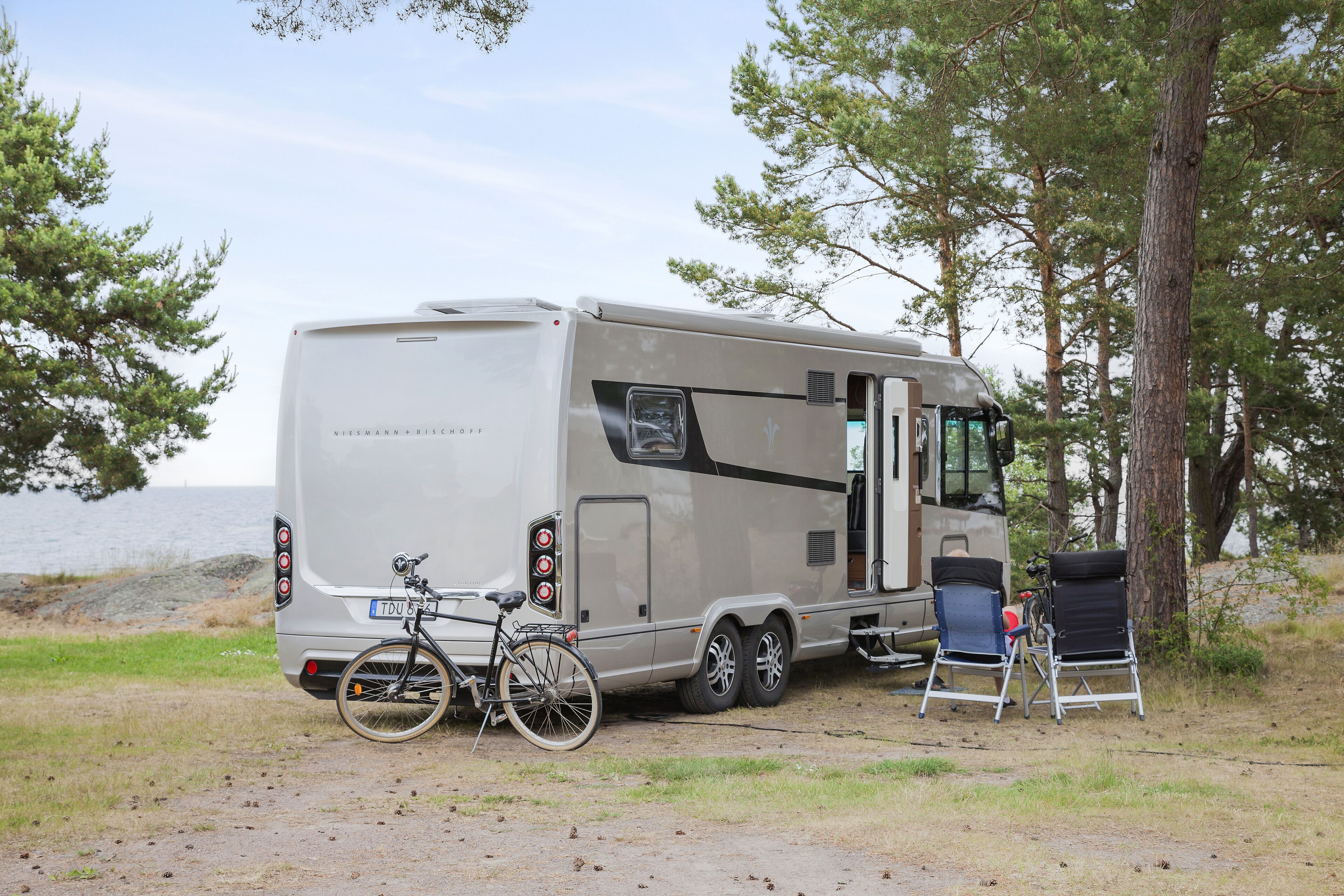 A recreational vehicle parked by a lake with a bicycle and chairs.