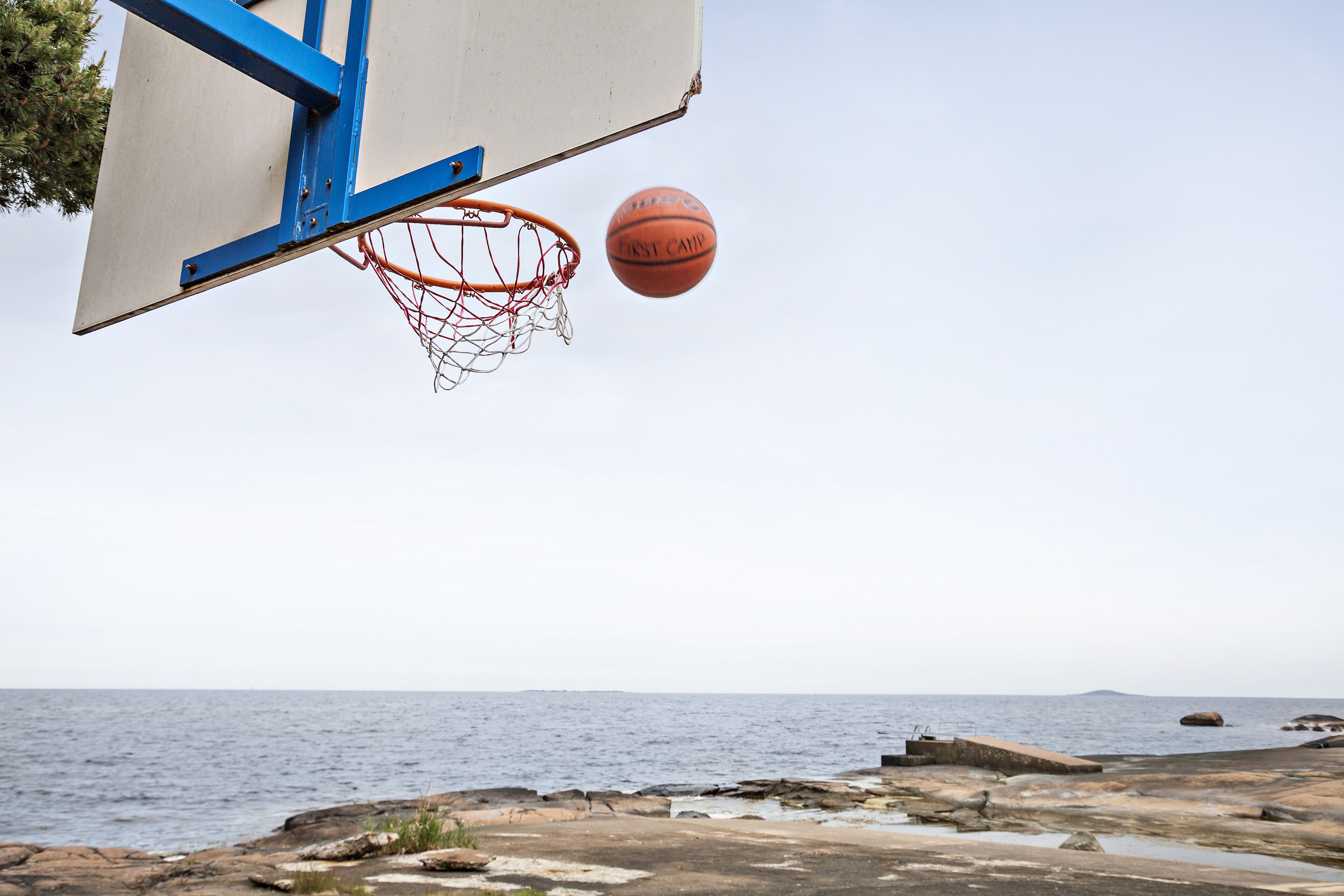 A basketball hoop with a net and a basketball in mid-air, overlooking a rocky shoreline with the sea in the background.