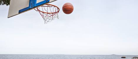 A basketball hoop with a net and a basketball in mid-air, overlooking a rocky shoreline with the sea in the background.