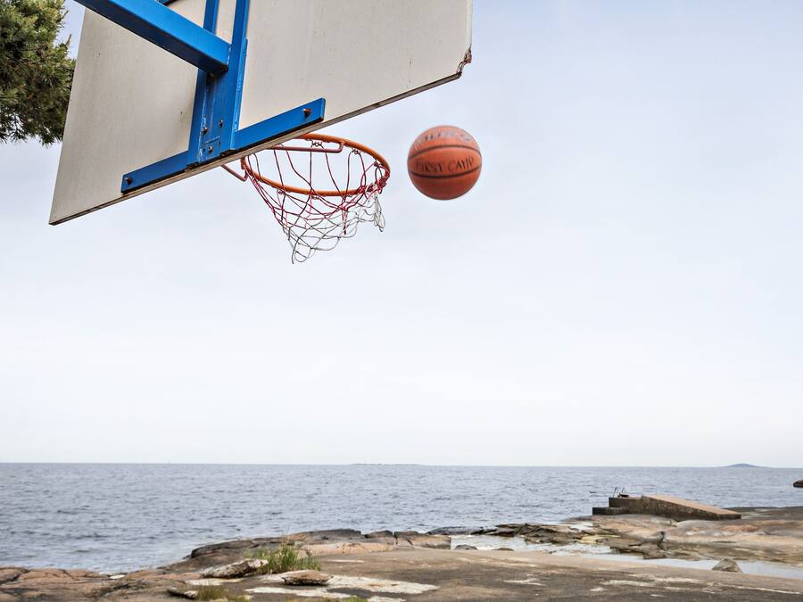 A basketball hoop with a net and a basketball in mid-air, overlooking a rocky shoreline with the sea in the background.