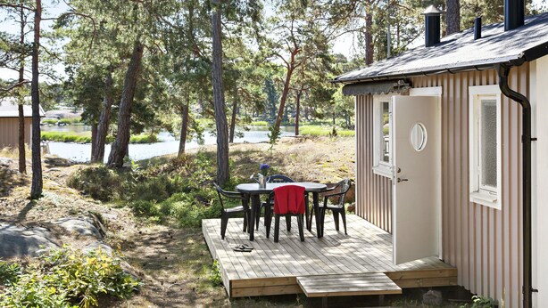 A small cabin with a deck, a table with chairs, and a view of a lake surrounded by trees.