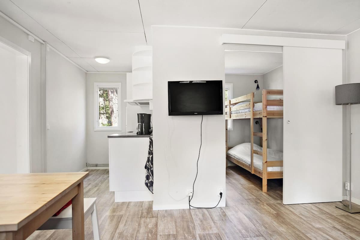 Una habitación moderna y compacta con una cama litera de madera, un televisor de pantalla plana en la pared, una pequeña cocina y una mesa de comedor.