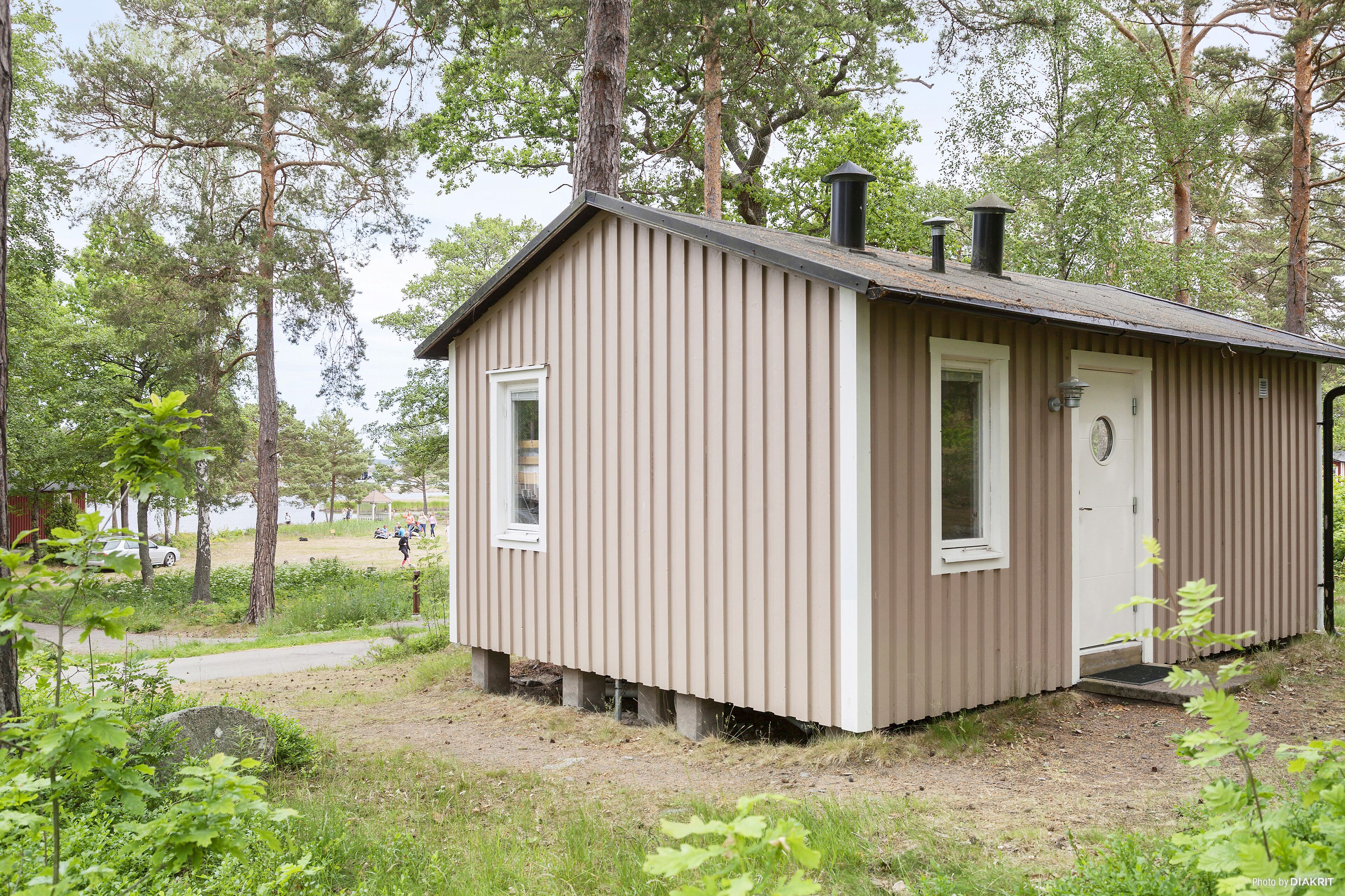 A small, single-story cabin with a sloped roof and a chimney, surrounded by trees and a dirt path.