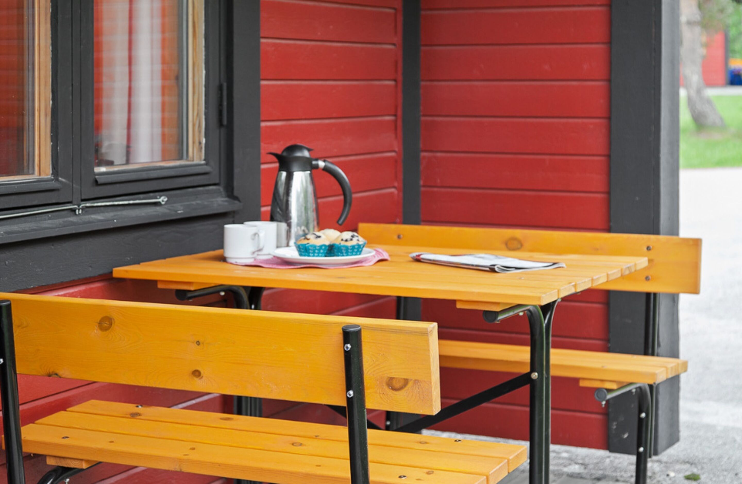 A wooden table with a metal chair, a cup, a plate with a pastry, and a coffee pot.