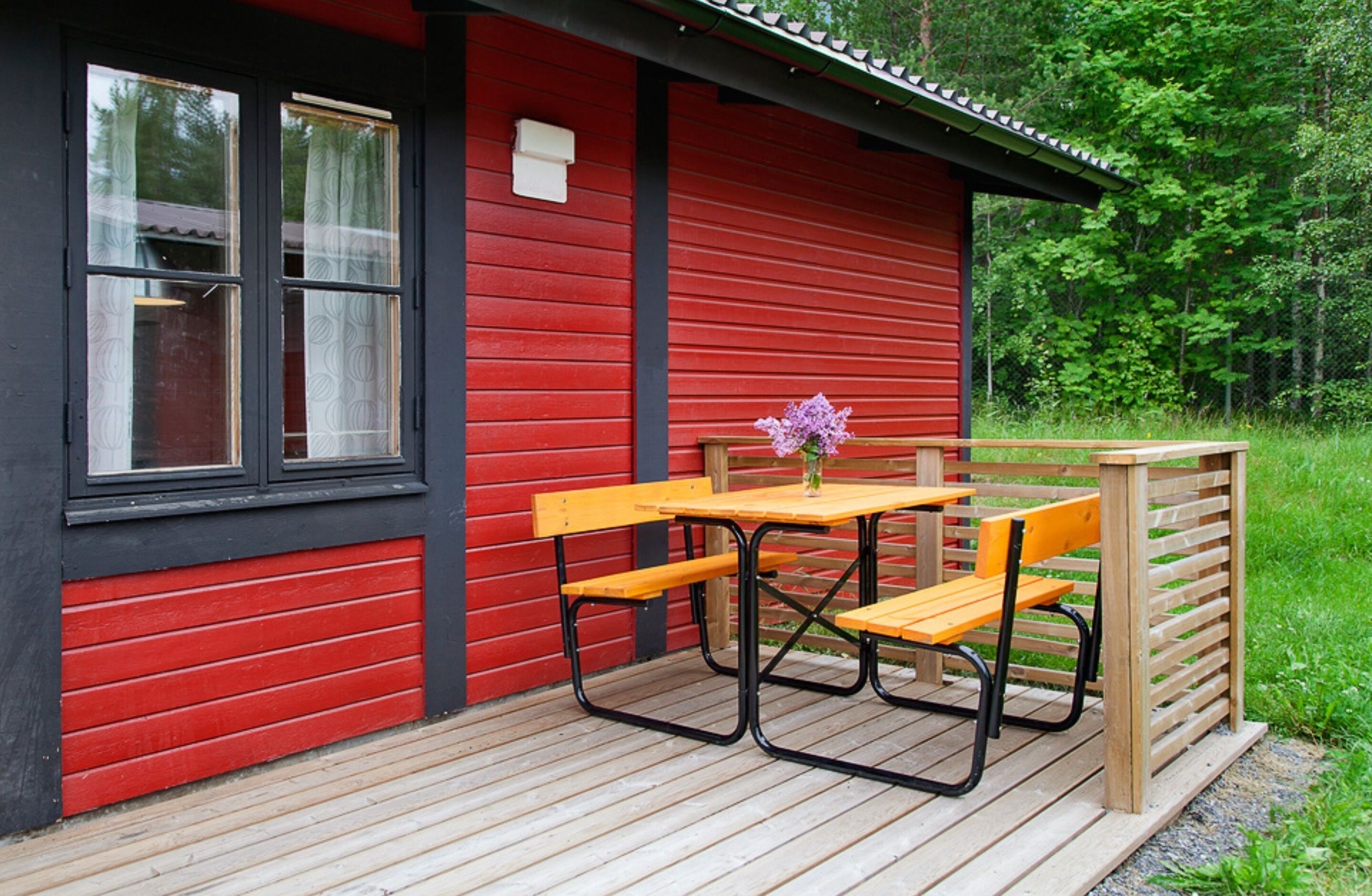 Una cabaña roja de madera con un porche de madera, una mesa de picnic y una banca.