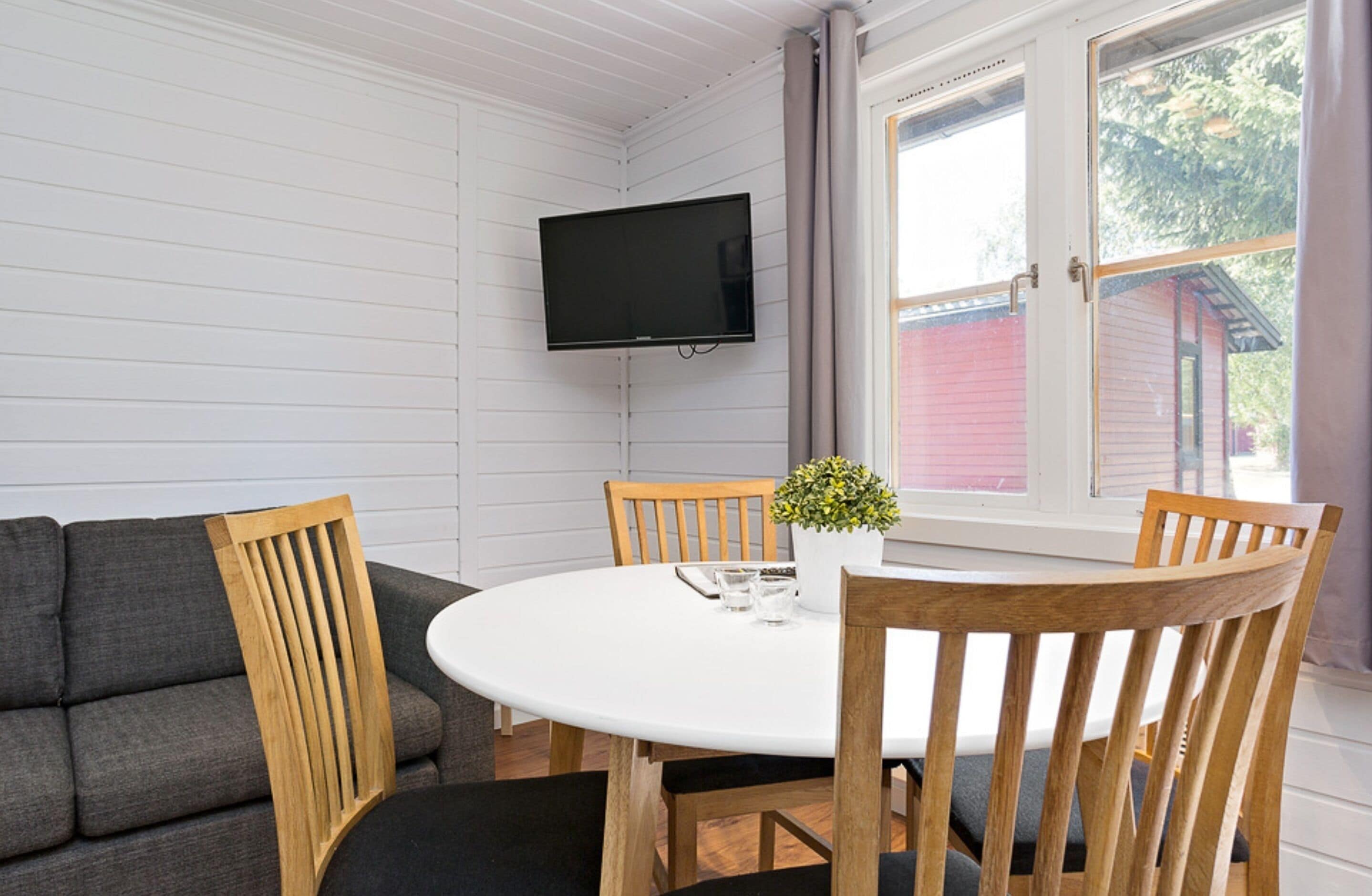 A room with a white table and four wooden chairs, a grey sofa, a flat-screen TV mounted on the wall, and a window with curtains.