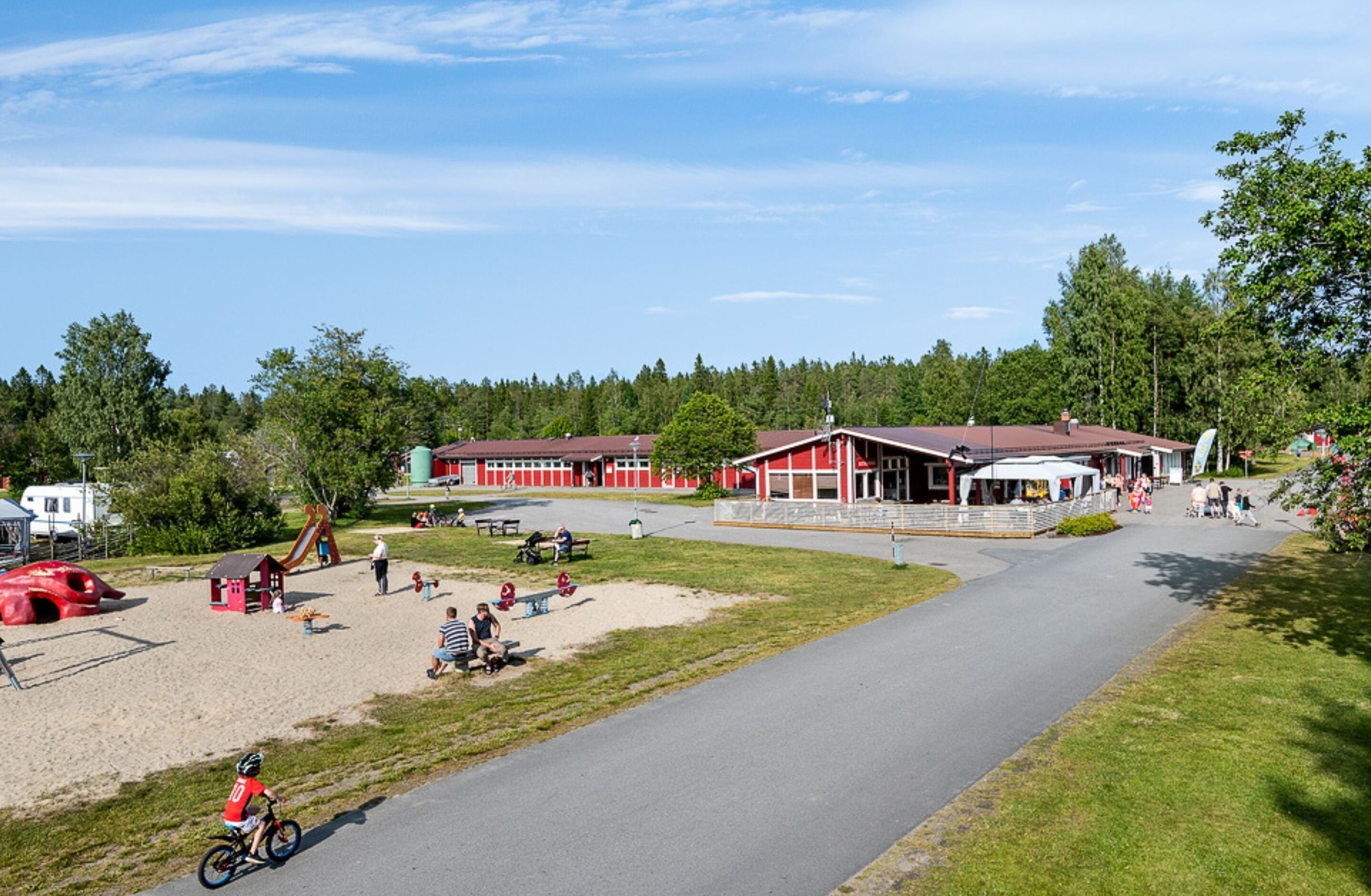 A park with playground equipment, a red building with a covered area, and a paved path.