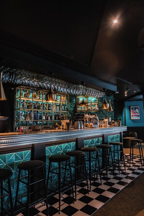 Eine Bar mit Schachbrettmuster-Boden, Barhocker und einem gut beleuchteten Kamin mit Flaschen und Gläsern.