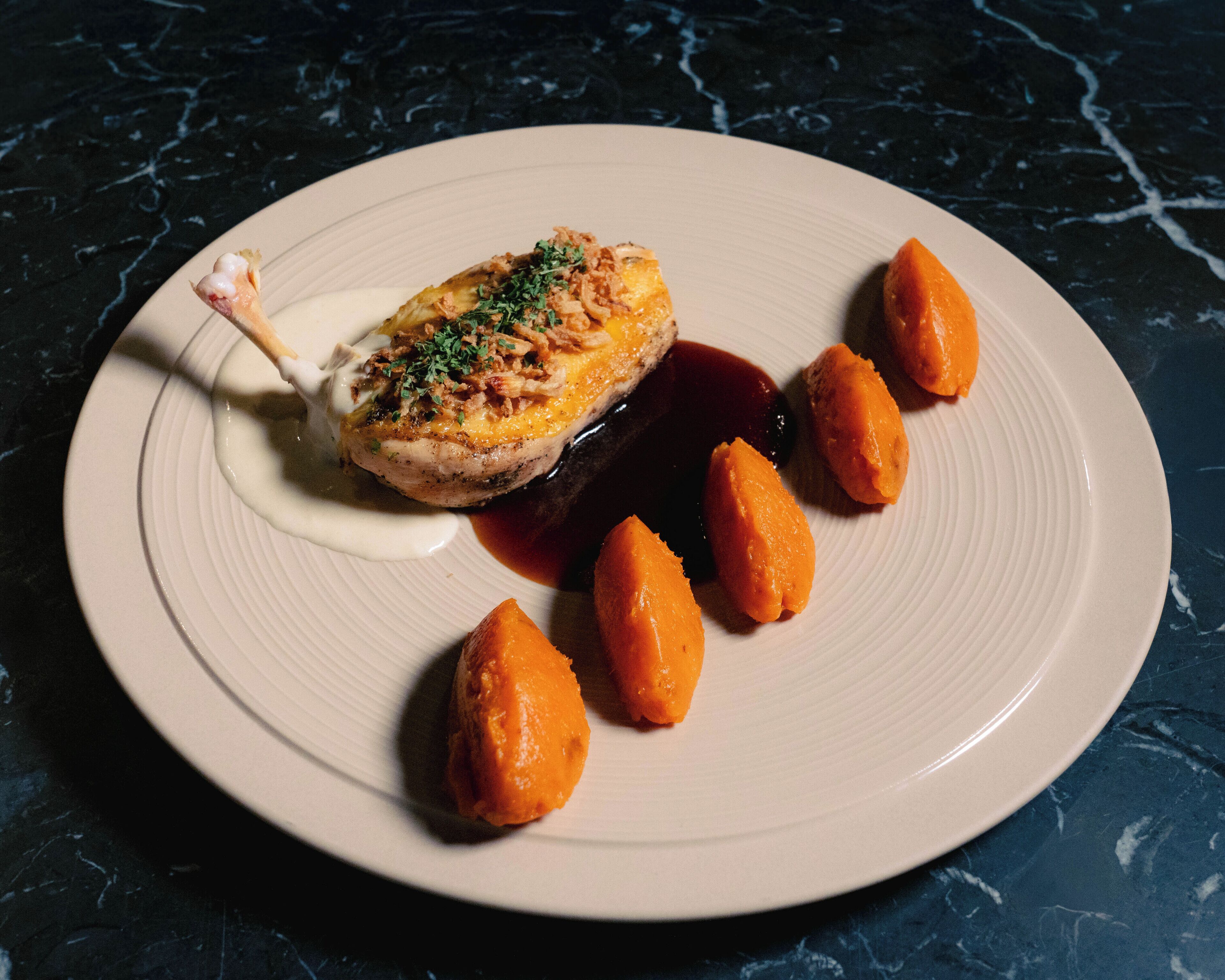 Un plato con un trozo de carne cubierto de queso y hierbas, acompañado de salsa oscura y varios elementos con forma de zanahoria.