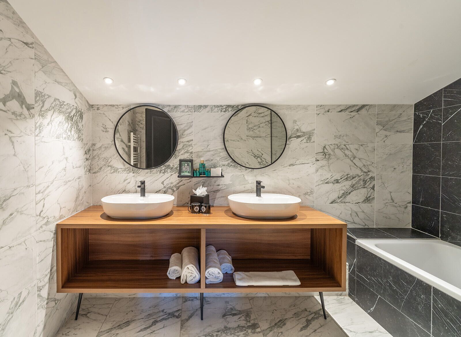 A modern bathroom with two sinks, wooden vanity, marble walls, and a bathtub.