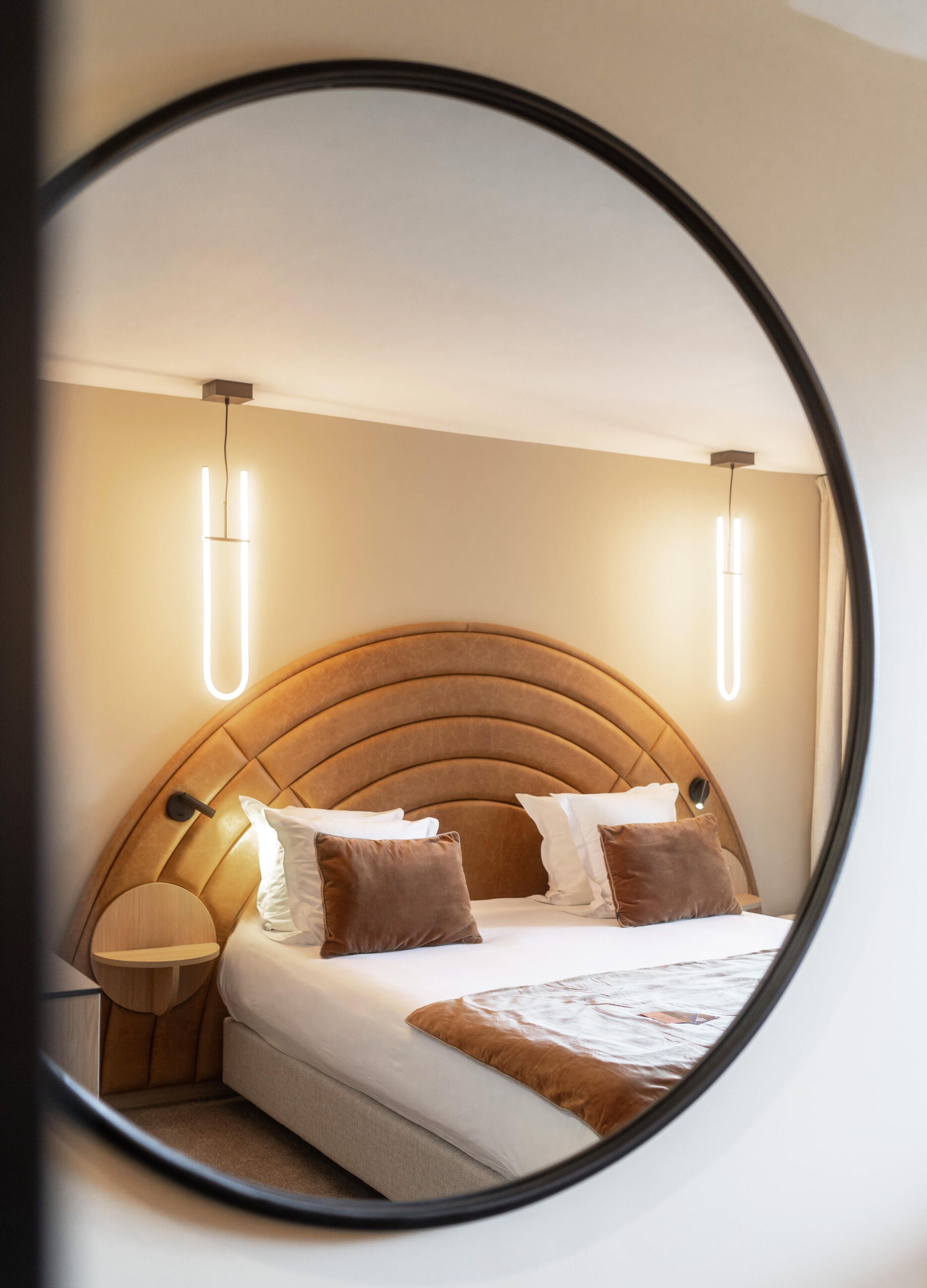 A circular mirror reflecting a neatly made bed with a wooden headboard, two bedside tables, and wall-mounted lights.