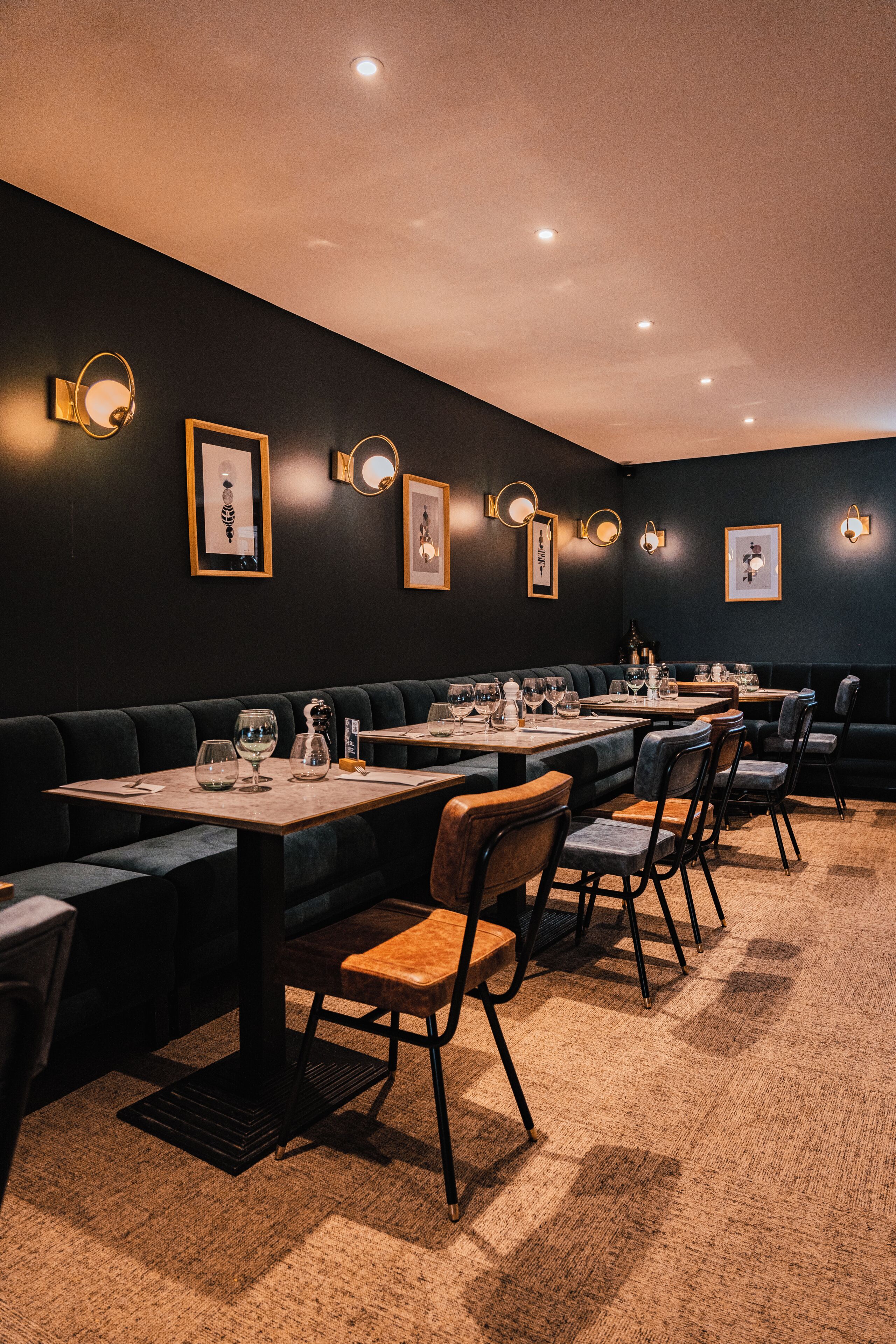 Un restaurante con paredes oscuras, apliques redondos en la pared y suelo alfombrado. Las mesas están dispuestas con vasos y cubiertos.