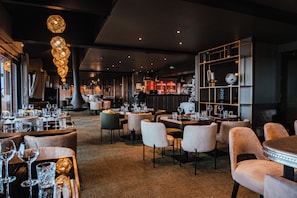 Ein Restaurantinneres mit mehreren Tischen, Stühlen, einer Bar und hängenden Pendelleuchten.