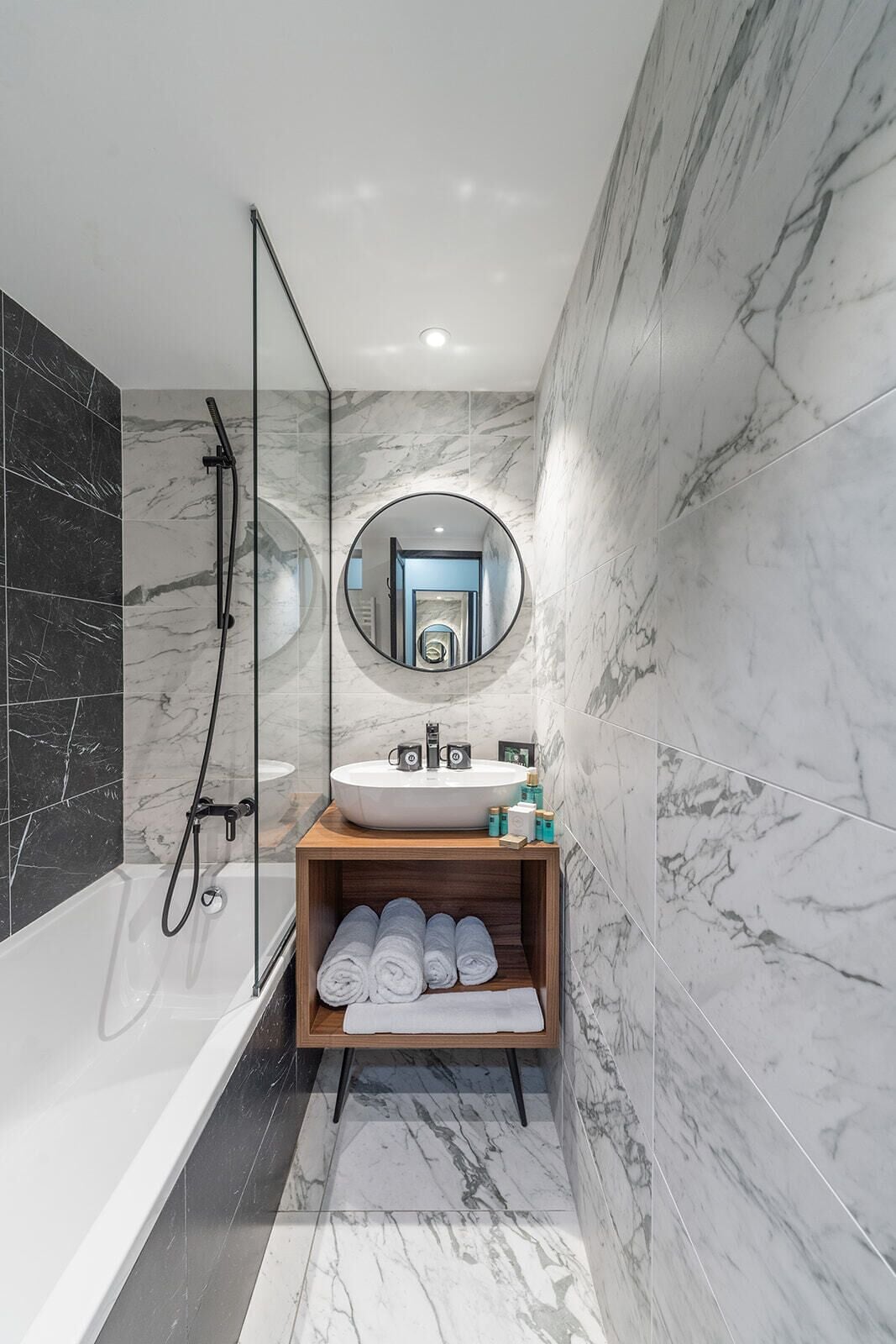 A modern bathroom with a bathtub, a round mirror, a sink, and a shelf with towels.