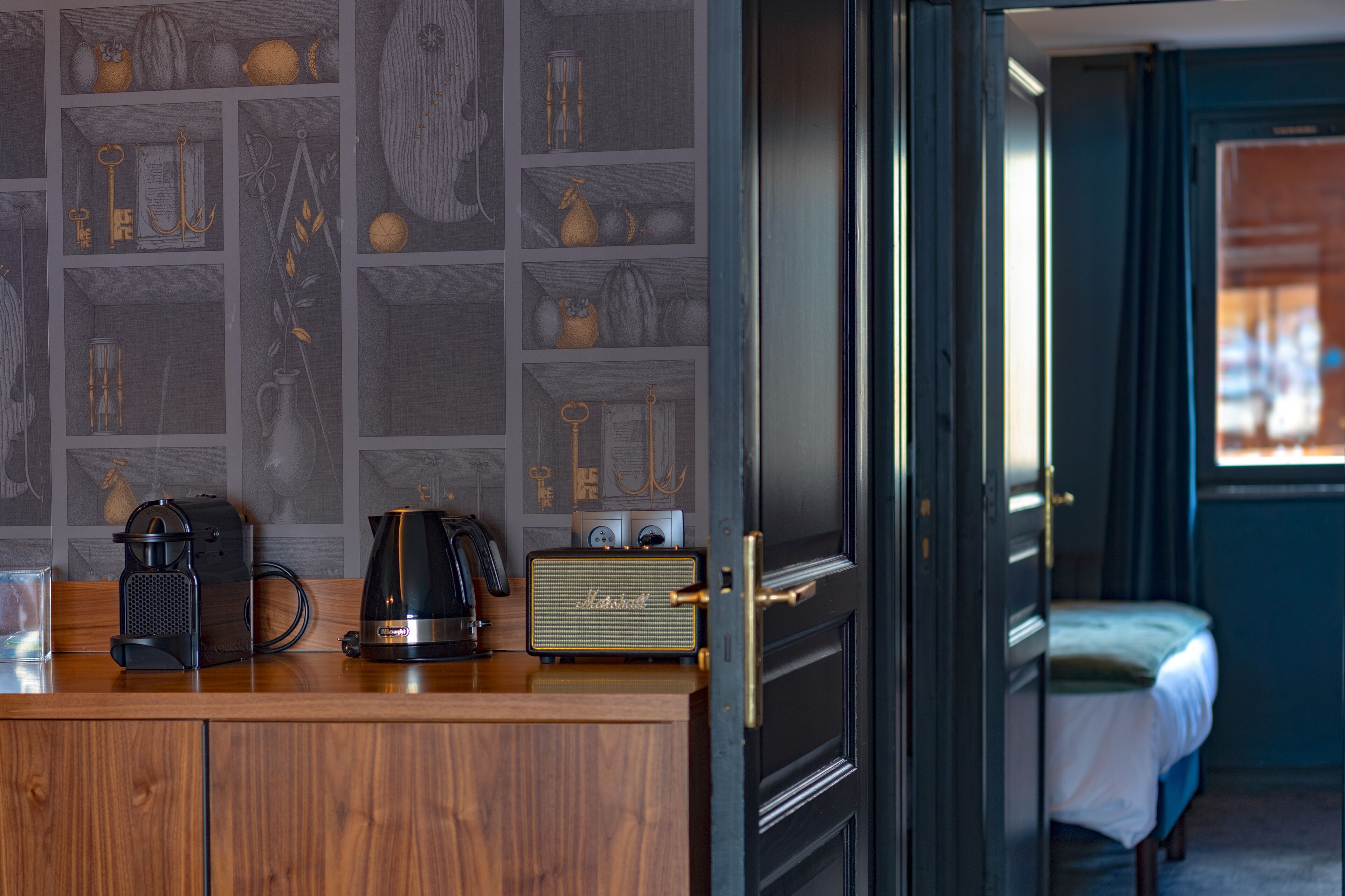 Una encimera de madera con una cafetera, un hervidor y una radio. Una puerta da a un dormitorio con una cama y una ventana.