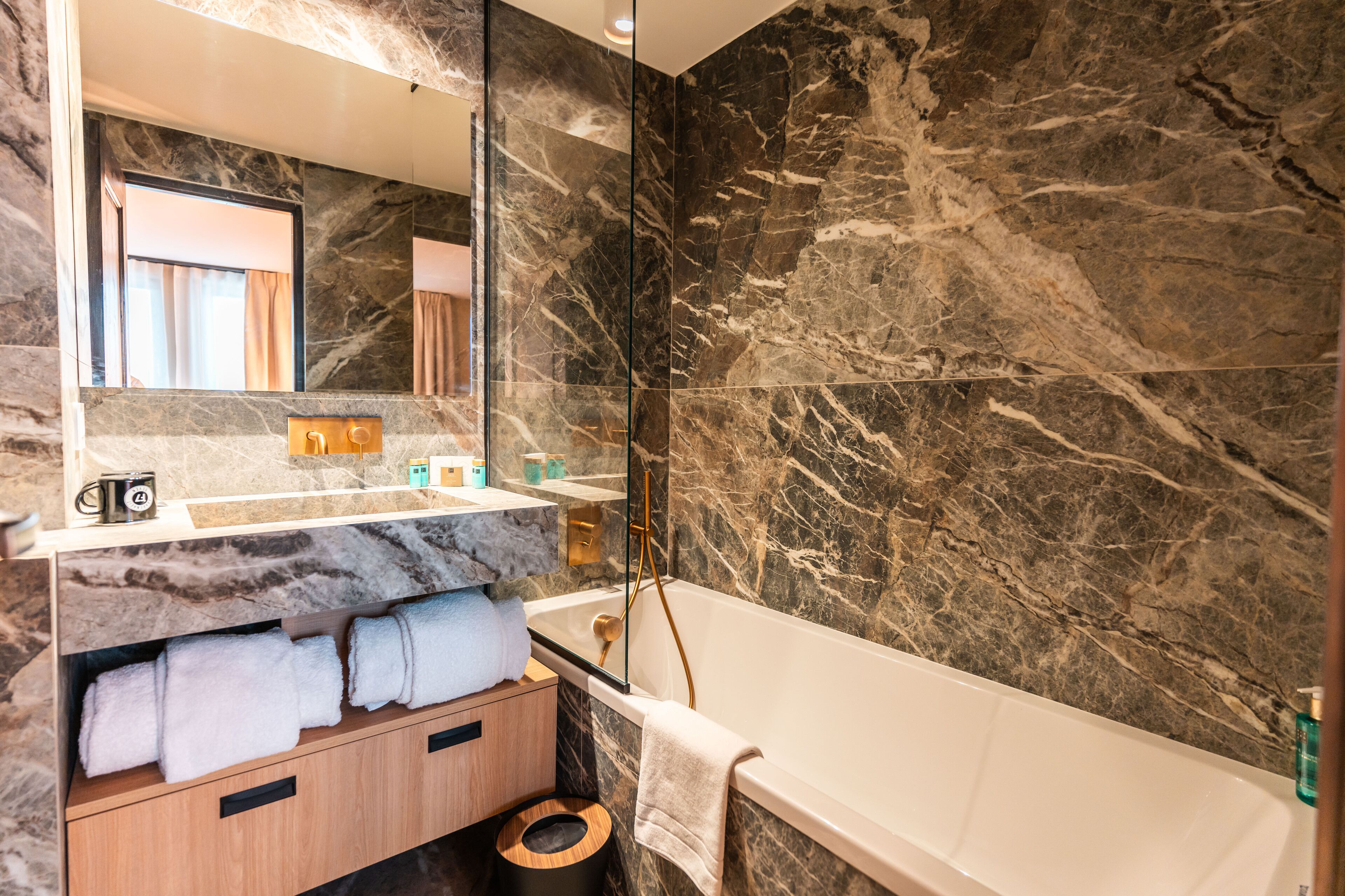 A modern bathroom with a marble wall, a large mirror, a bathtub, and a wooden vanity with towels.