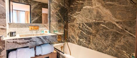 A modern bathroom with a marble wall, a large mirror, a bathtub, and a wooden vanity with towels.