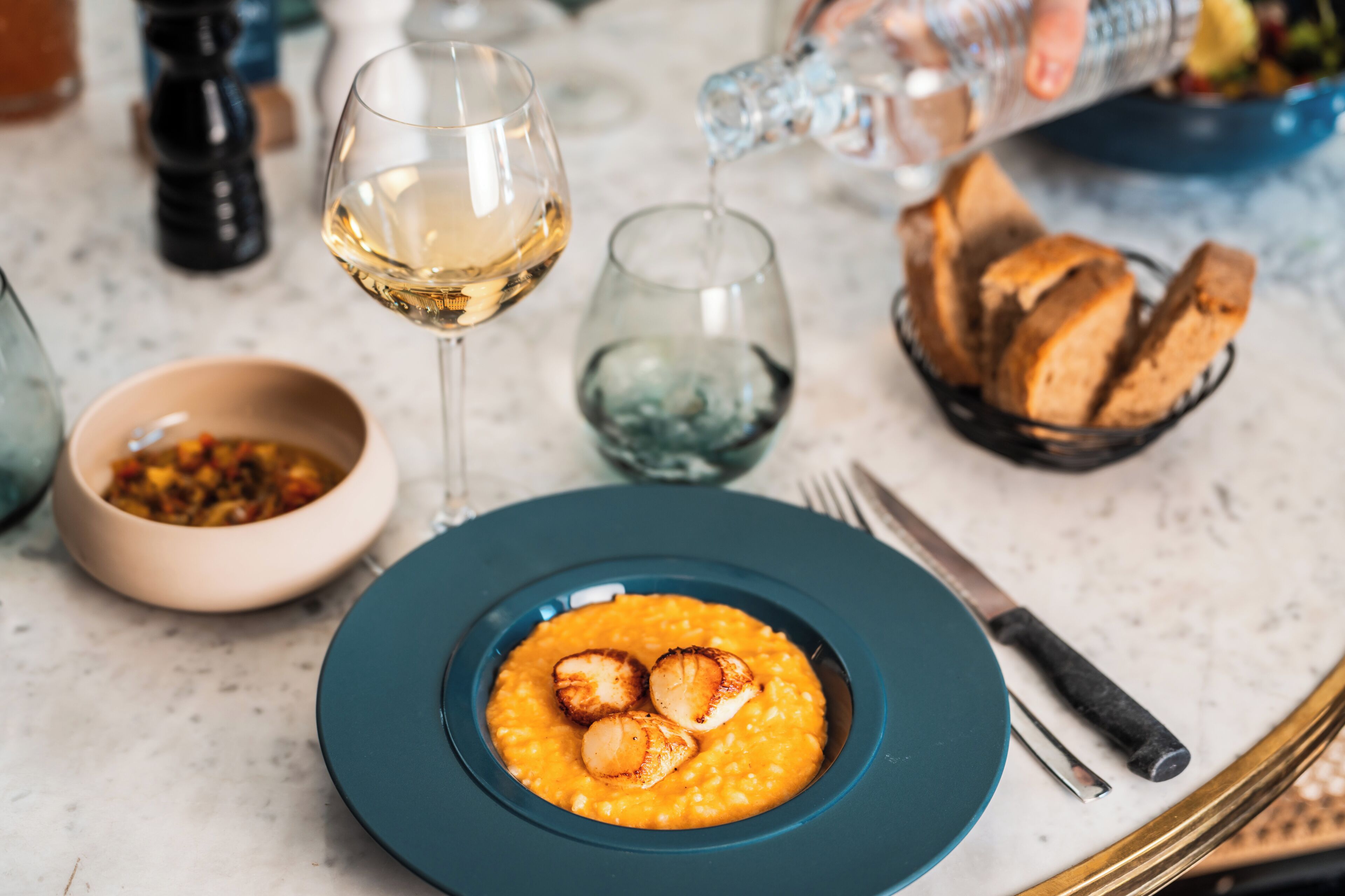 A table with a plate of food, a glass of white wine, a bowl of soup, and a basket of bread.