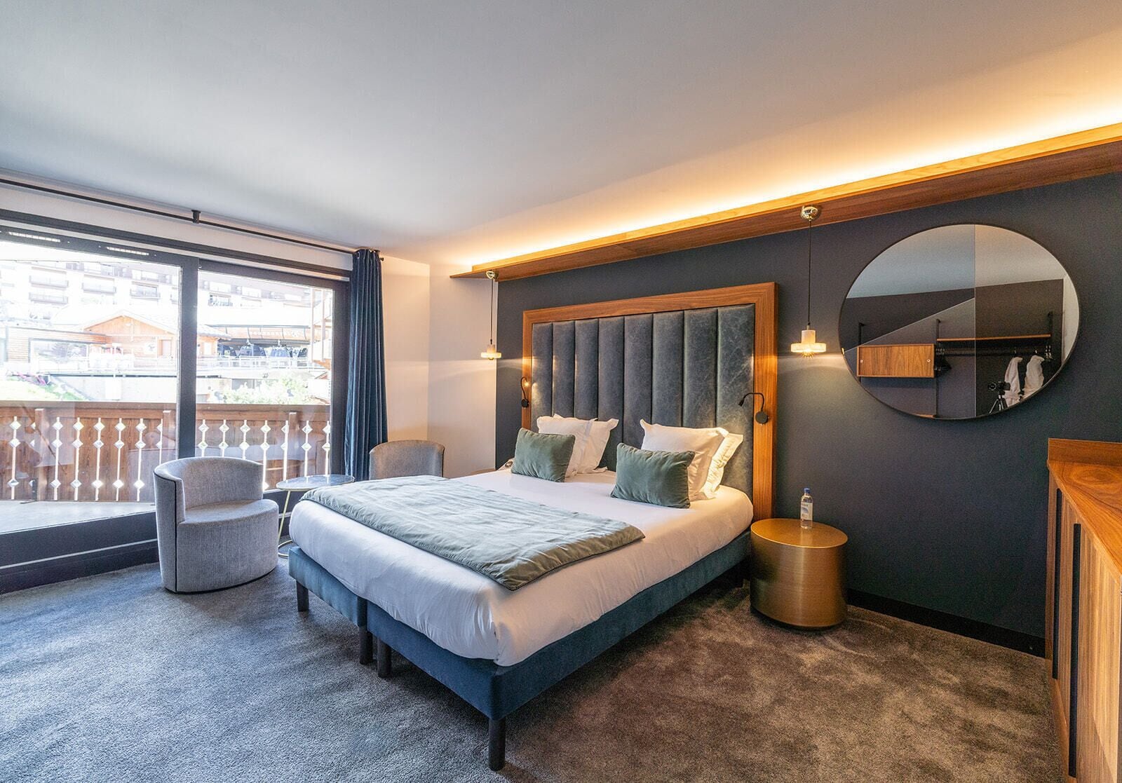 A modern hotel room with a large bed, a round mirror, a small table, and a balcony with a view.