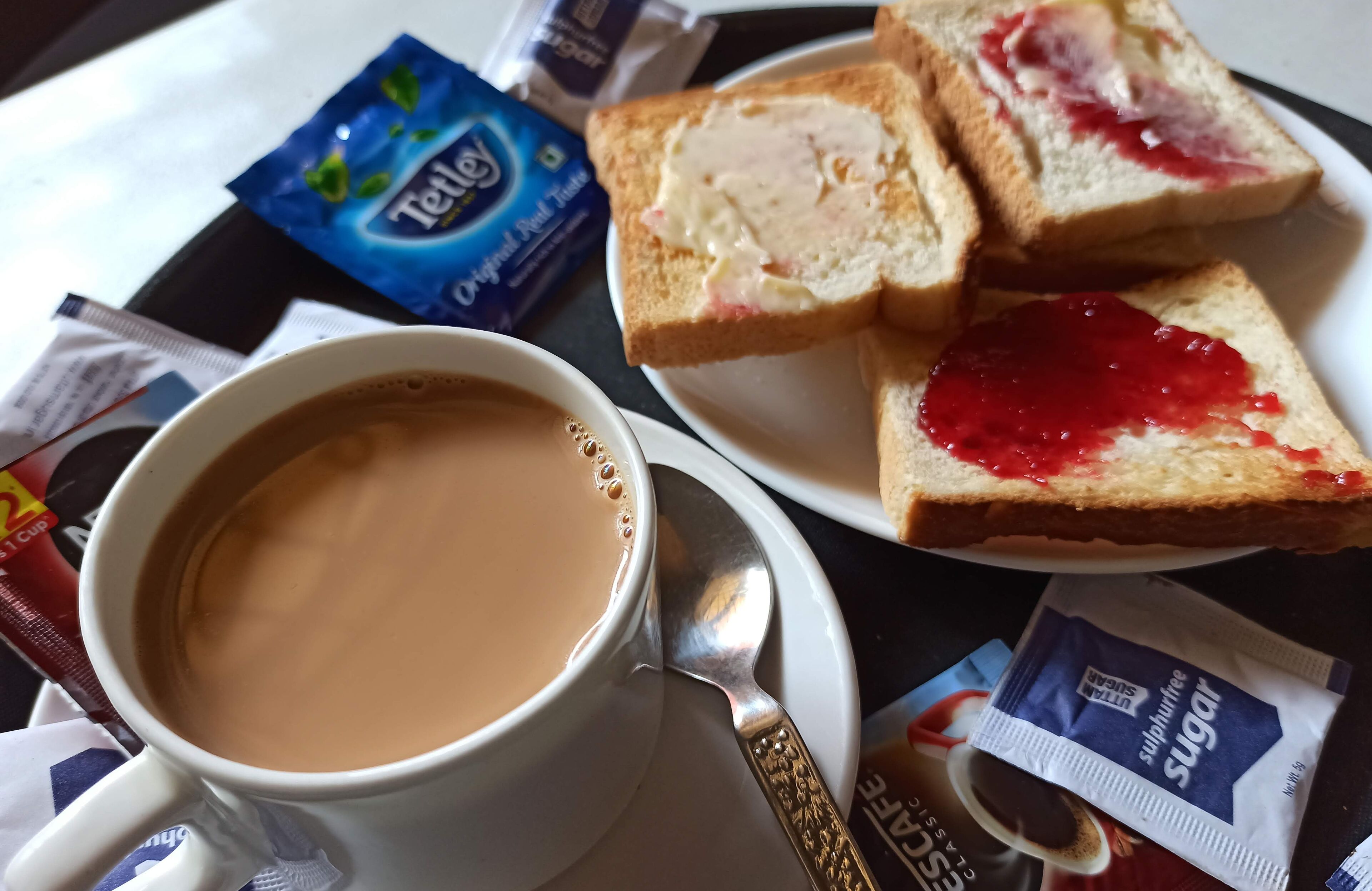 Eine Tasse Kaffee mit Löffel, ein Teller mit zwei Scheiben Brot mit Marmelade sowie Teebags und Zucker auf einem Tisch.