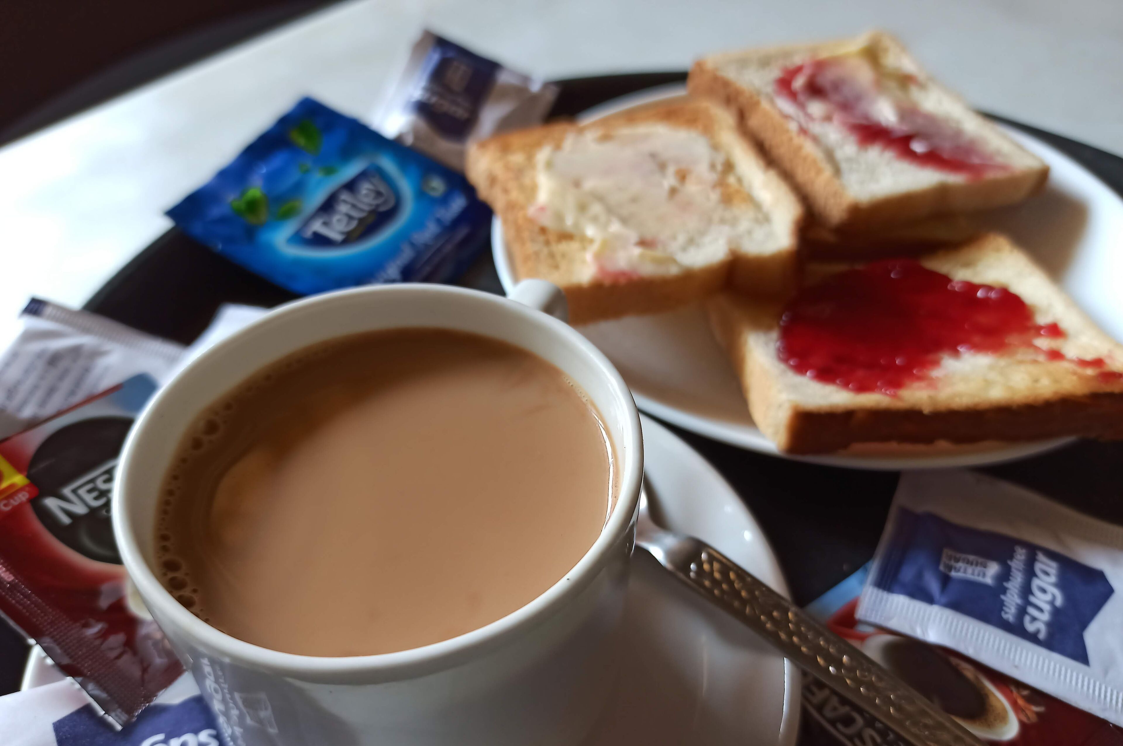 Una tazza di caffè, due fette di pane tostato con marmellata, una bustina di zucchero e una bustina di tè su un vassoio.