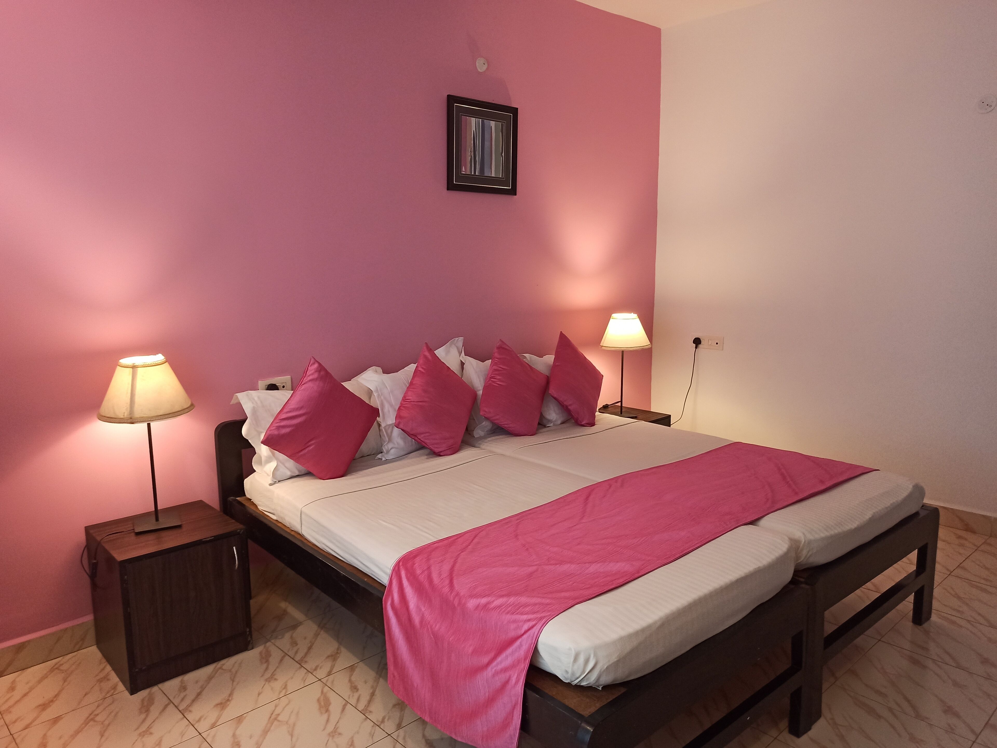 A bed with a pink blanket, white bedding, and pink pillows in a room with pink walls and a wooden headboard.