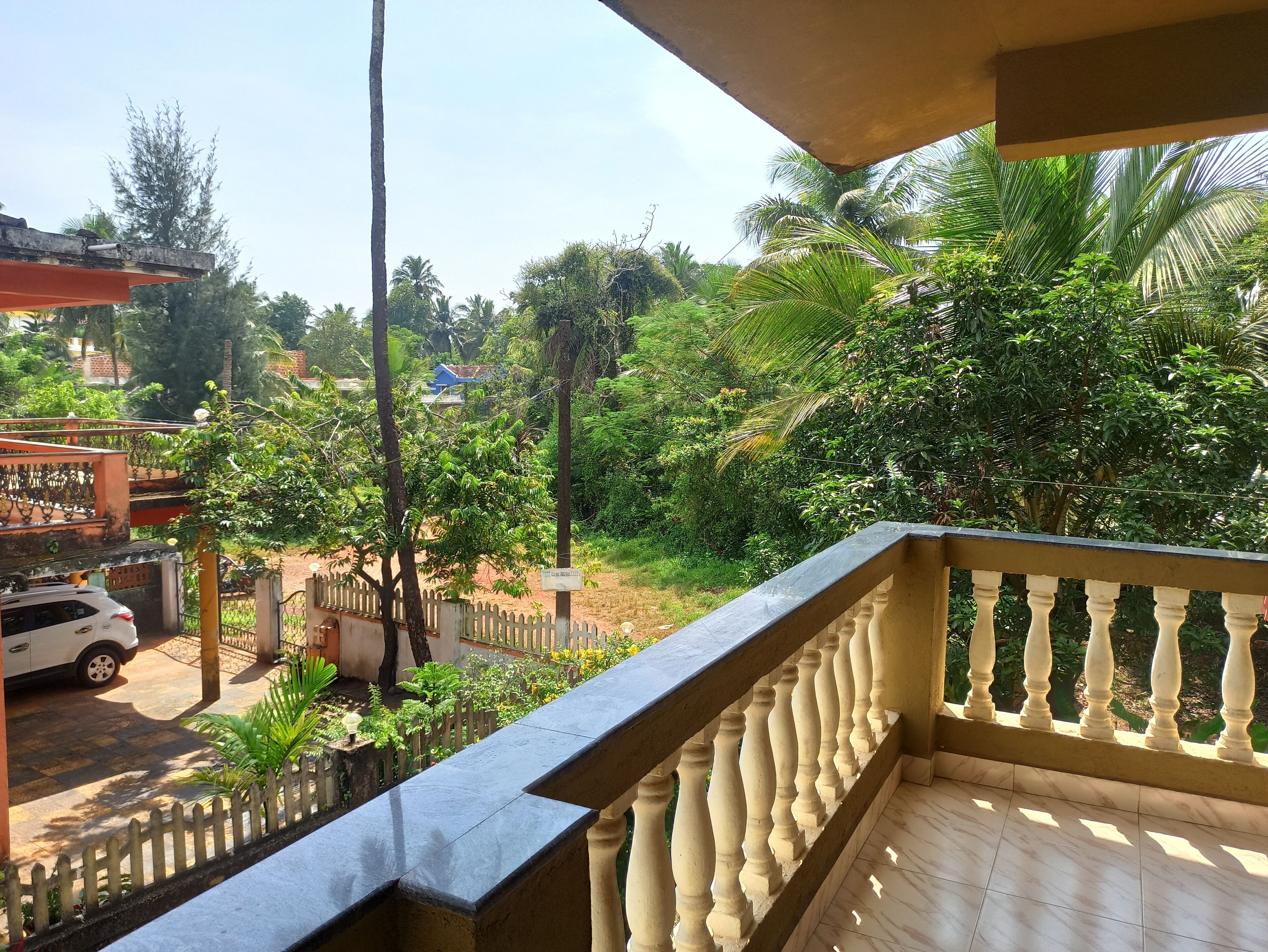 A balcony with a view of a garden, a white car parked outside, and a building in the background.