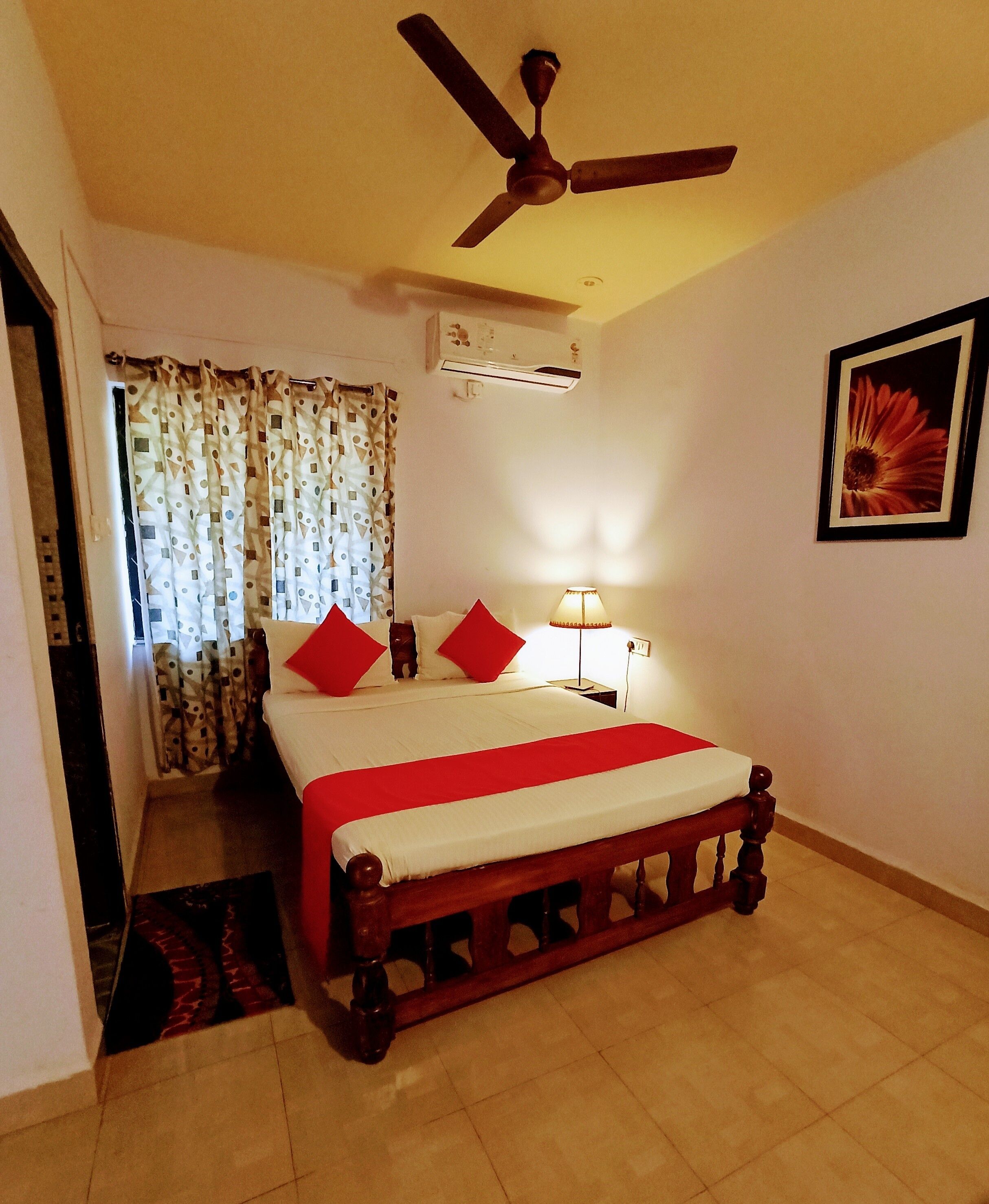 A bedroom with a bed, a ceiling fan, an air conditioner, a nightstand, a lamp, and a framed picture on the wall.
