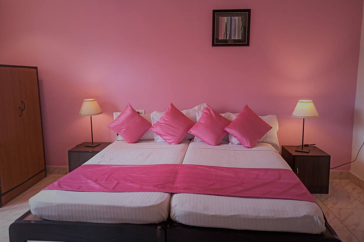 Een tweepersoonsbed met roze en witte beddengoed, twee bedlampjes en een ingelijst schilderij aan de muur.