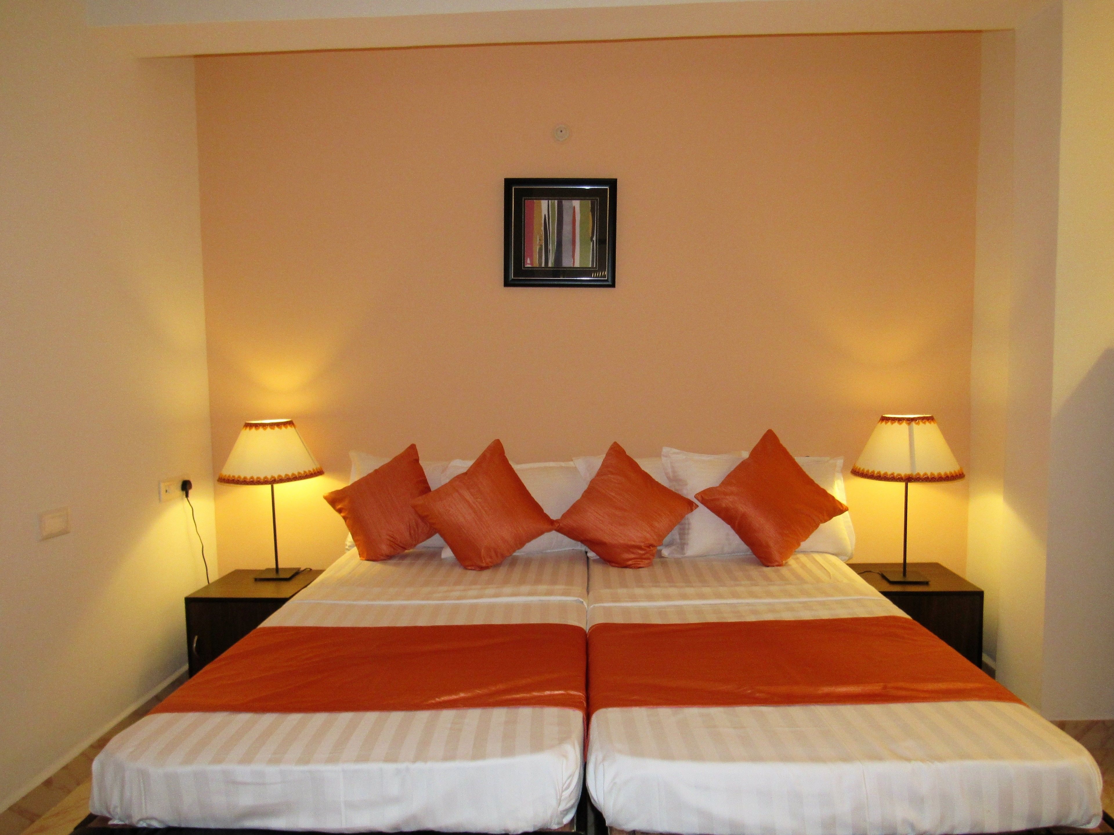 A double bed with orange bedding and pillows, two bedside lamps, and a framed picture on the wall.