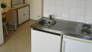 A compact kitchen with a sink, stove, and a small refrigerator. There is a wooden chair and a cabinet with a mirror.