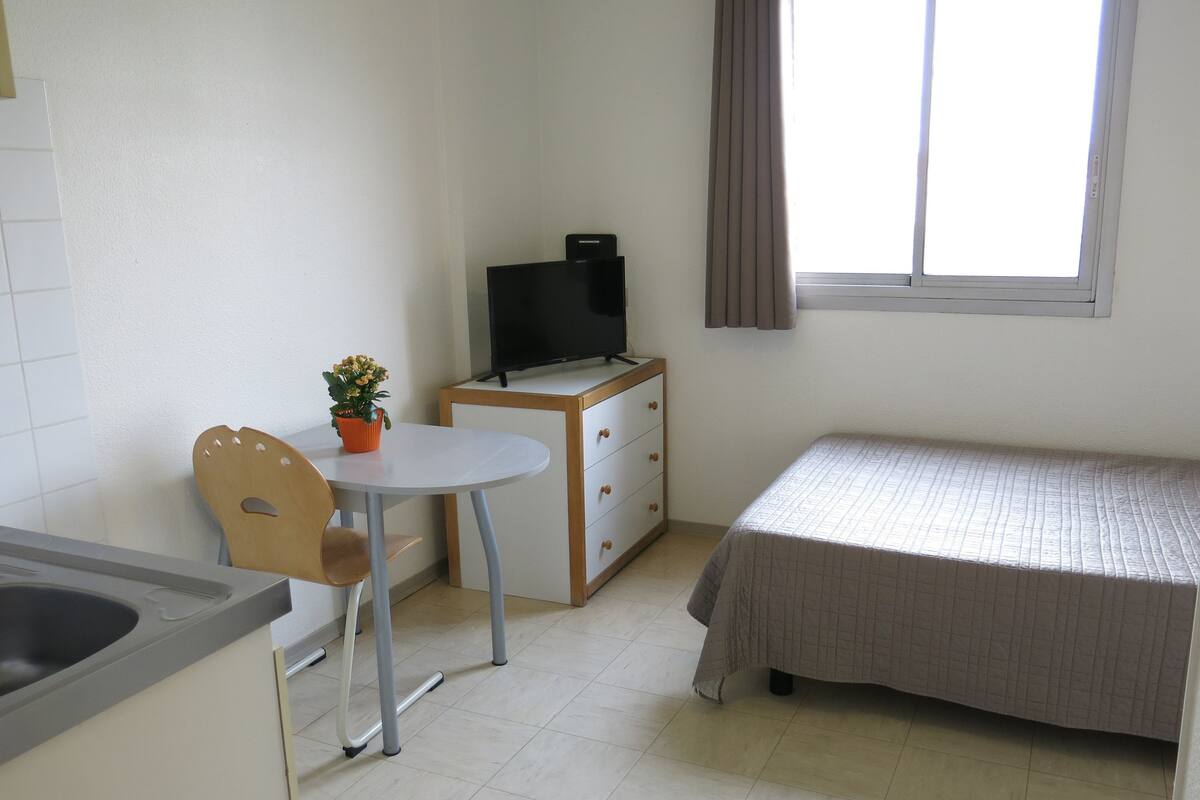 Una habitación pequeña y limpia con cama, escritorio, silla, televisión y ventana con cortinas.