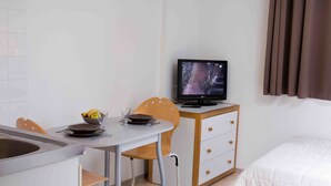 A compact hotel room with a bed, a small desk with a TV, a chair, and a dining area with a bowl of fruit and a glass.