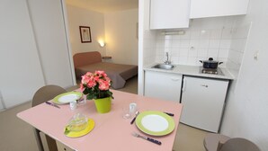 A compact hotel room with a dining area featuring a pink table set with plates, cutlery, and a vase of flowers. A bed and a small kitchenette with a refrigerator and sink are visible in the background. - ABITEL - Les Studines d'Ornon (Villenave-d'Ornon)