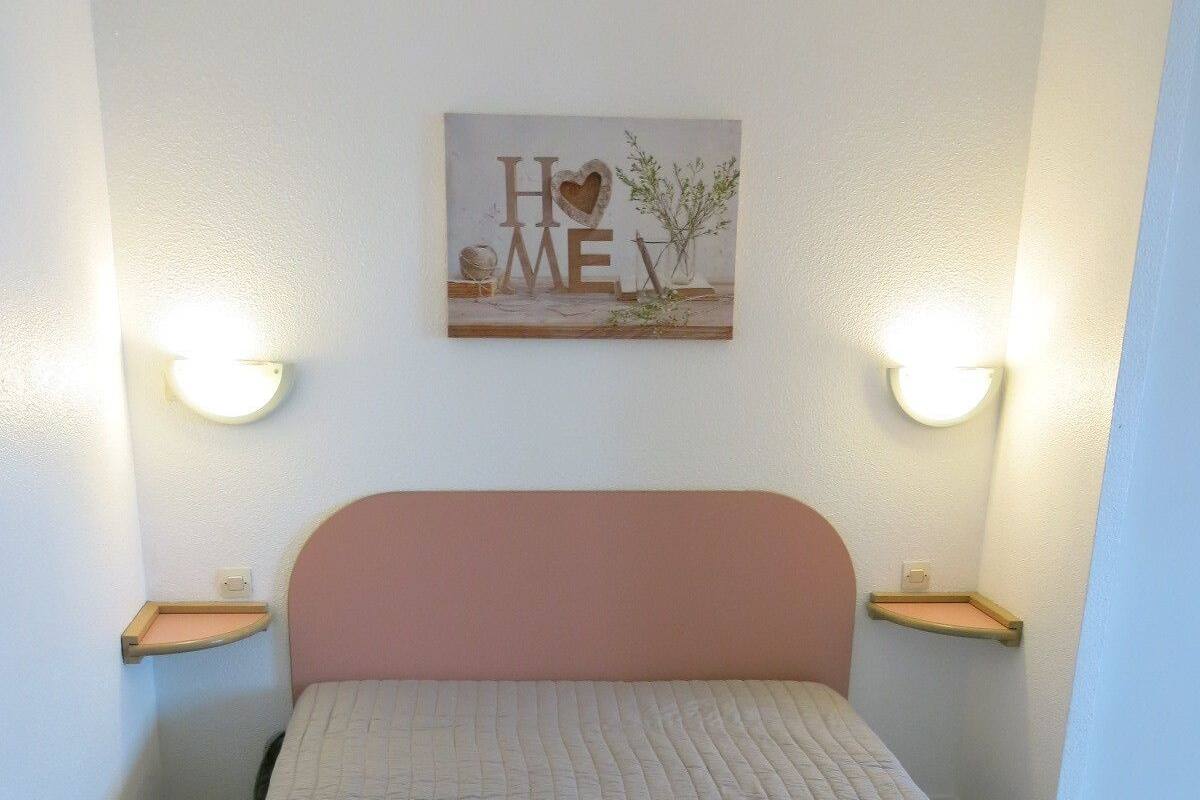 Un dormitorio con cama, cabecera, mesitas de noche y una lámpara fijada a la pared.