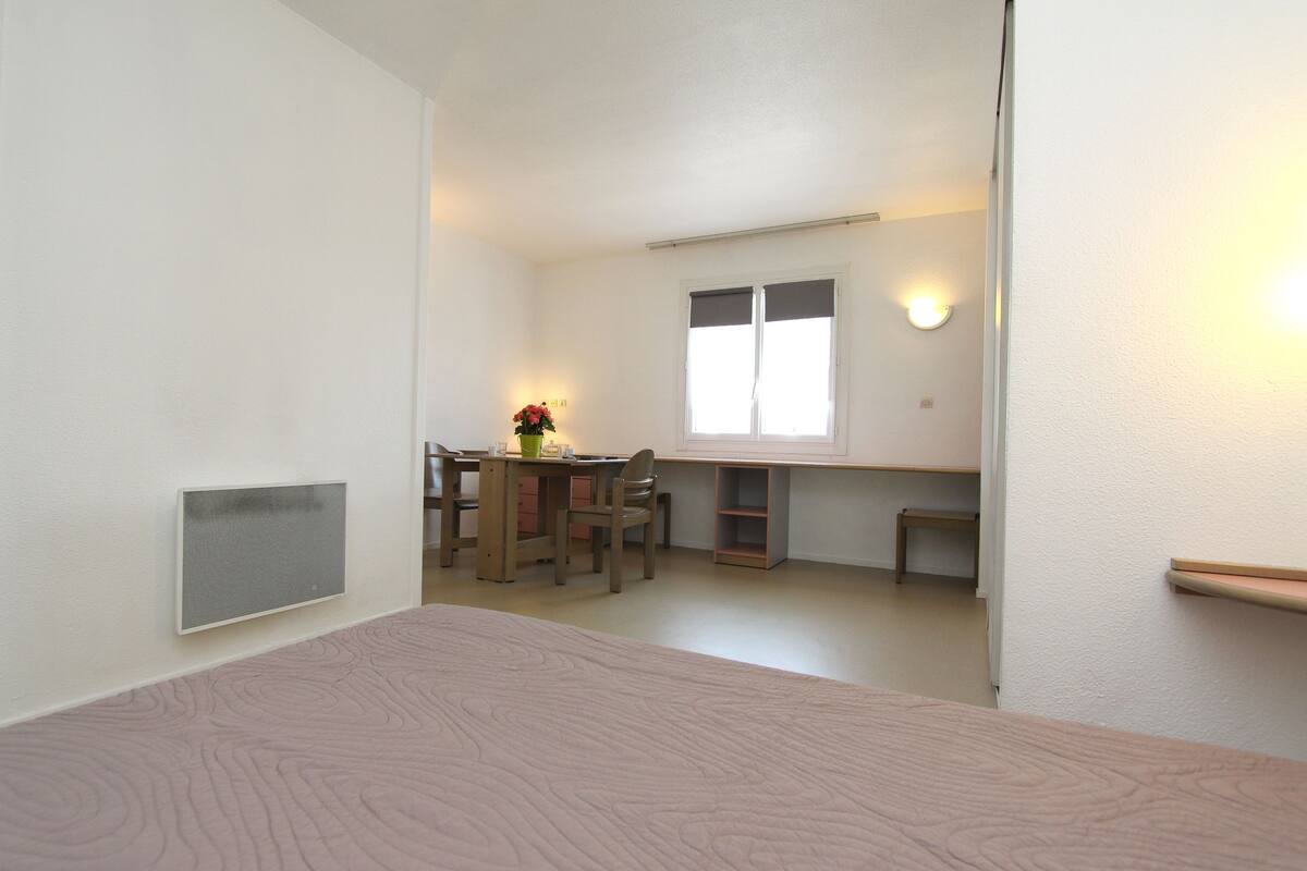 Una habitación pequeña y bien iluminada con una cama de madera, un escritorio con una silla y una ventana con persianas.