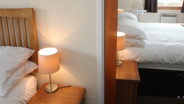 A hotel room with a wooden headboard, a bedside lamp, a bed with white linens, and a small wooden nightstand.