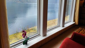 A room with a large window overlooking a lake and mountains. There is a red chair and a small potted orchid on the windowsill. - Visit Undredal (Aurland)