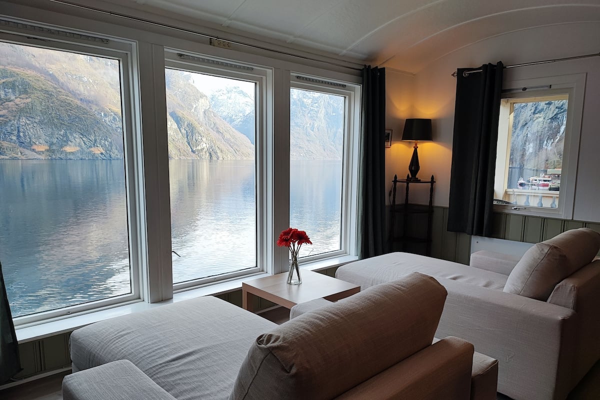 A modern room with large windows offering a view of a lake and mountains. There are two sofas and a small table with a vase of flowers.