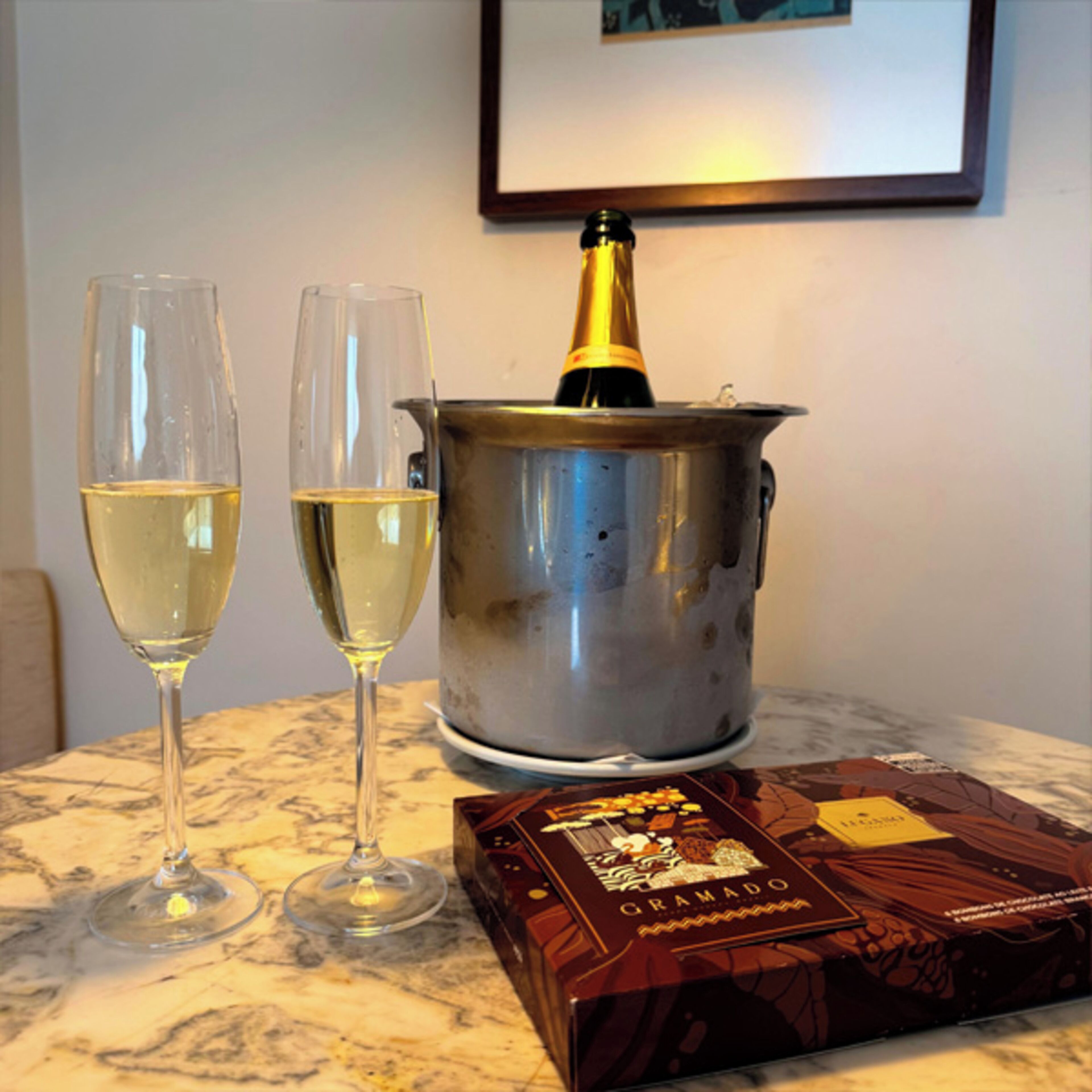 Dos copas de vino blanco, una botella de champán y una caja de bombones sobre una mesa de mármol.